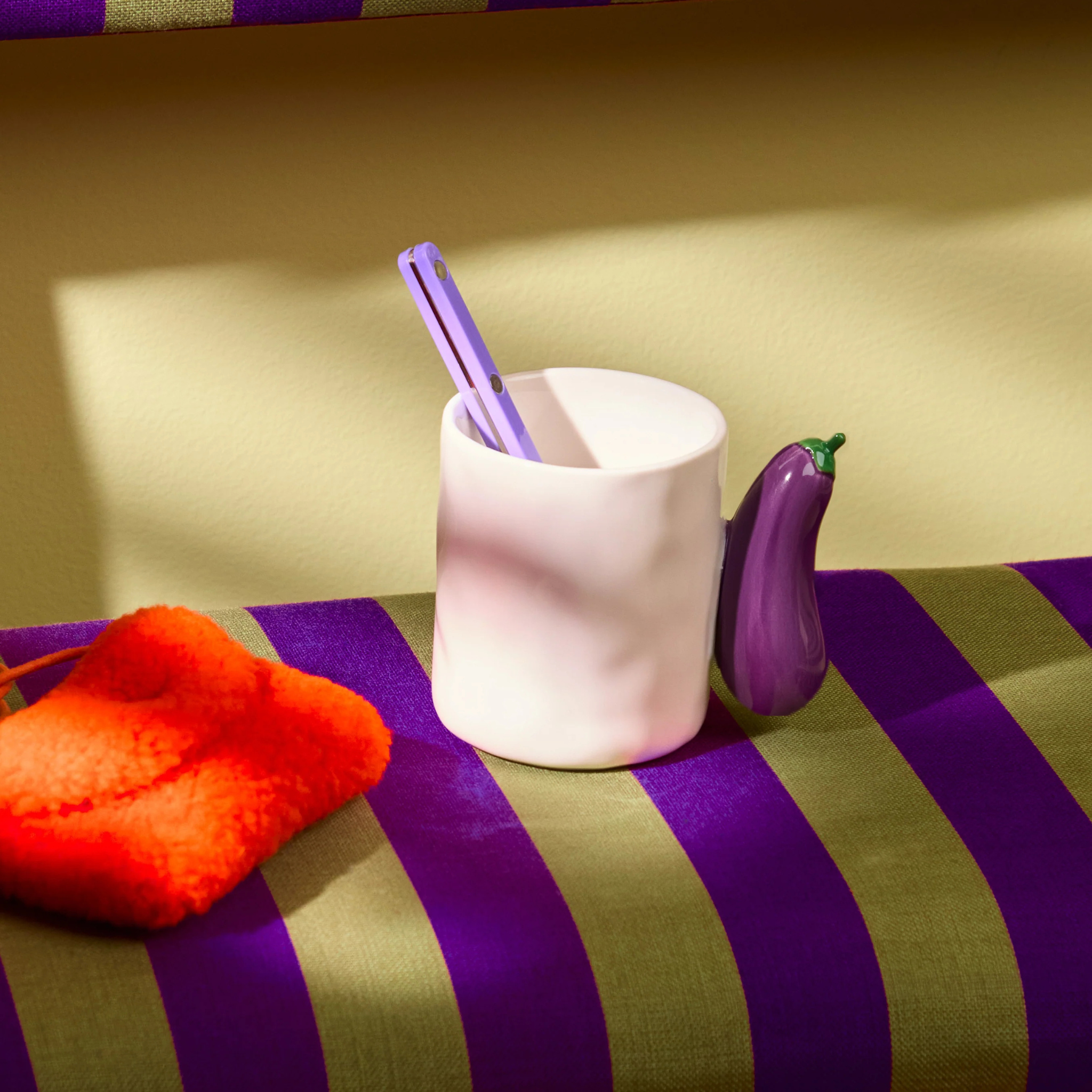 In de Fluid Market Mok Aubergine van Fluid Market, een witte mok van keramiek met een klein auberginevormig handvat, passen twee paarse pennen. Hij staat op een groen en paars gestreept oppervlak in de buurt van een oranje donzig object in zacht zonlicht.