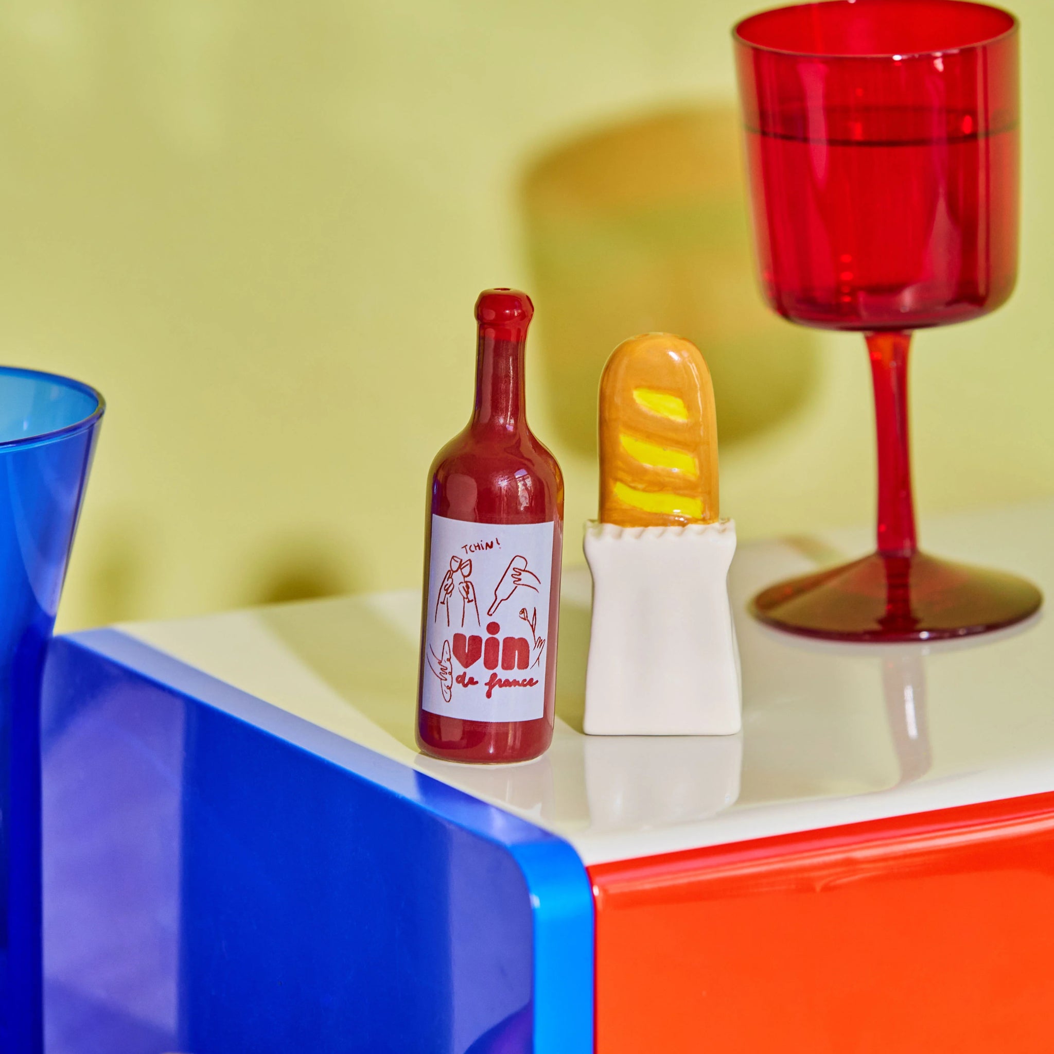 De Fluid Market Zout- en Pepervaatjes Baguette 2 st. van Fluid Market staat op een kleurrijk oppervlak met rode en blauwe glazen voor een vage gele achtergrond-een charmant tafelaccent voor elke omgeving.