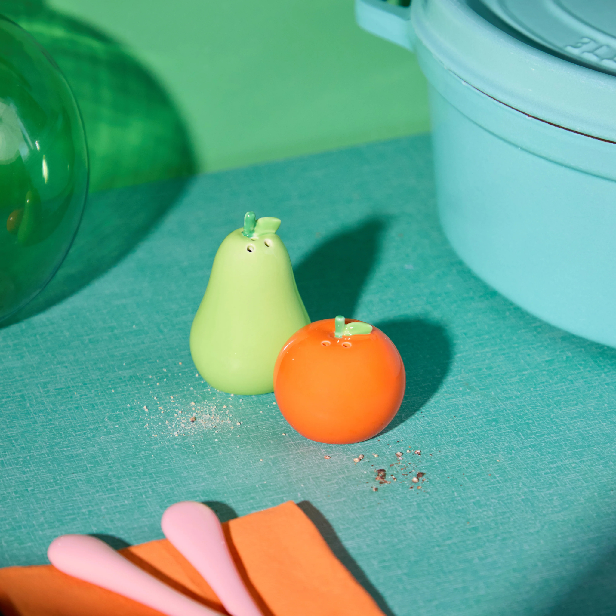 Een groene peervormige en oranje ronde Fluid Market Zout- en Pepervaatjes Appelsien Peer 2 st. met gezichtjes zitten op een groenblauwe ondergrond bij een blauwe pot en drie pastelkleurige lepels, met korrels verspreid tussen de shakers.