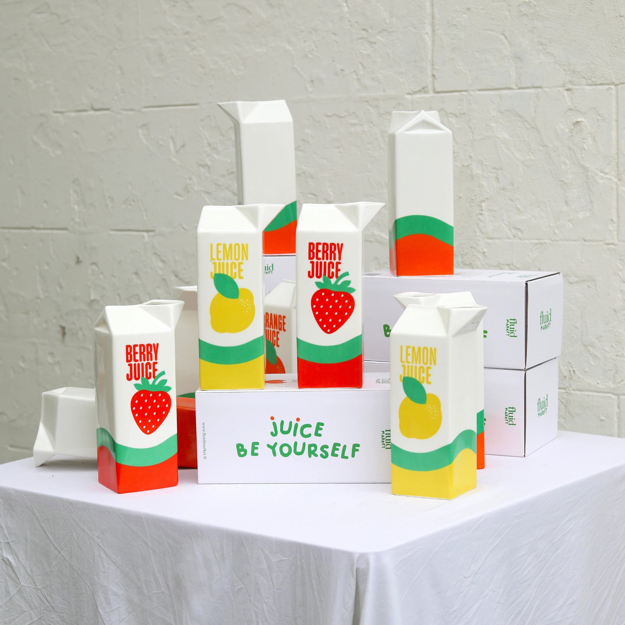 Fluid Market Citroensap Vaas dozen, met kleurrijke, op Frans design geïnspireerde minimalistische patronen, staan uitgestald op een wit tafelkleed. Daarachter staan eenvoudige witte Fluid Market-kartons en sappakken bij een witte muur met structuur.