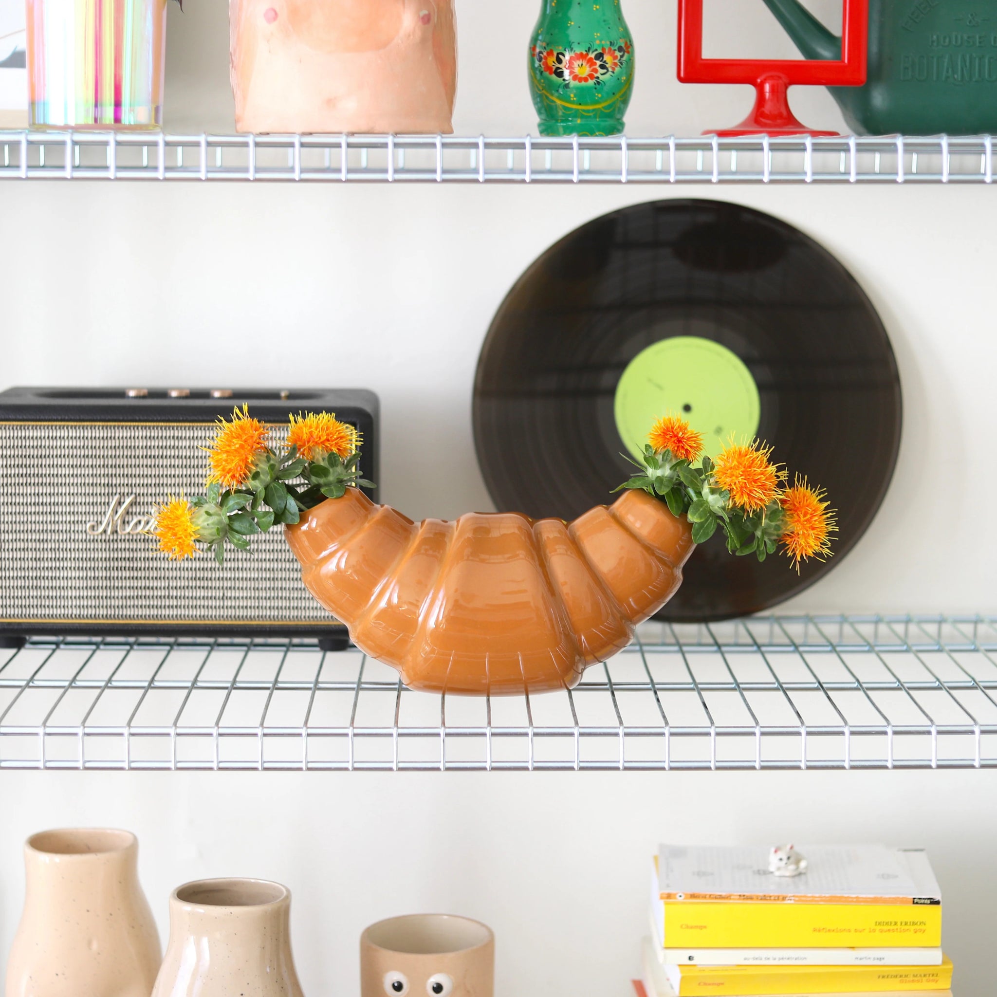 De Fluid Market Croissant Vaas van Fluid Market, een vaas van keramiek in de vorm van een croissant, toont oranje bloemen op een draadplank met kleurrijke decoraties, vinylplaten, luidsprekers en boeken - ideaal voor een speels interieur met Franse invloeden.