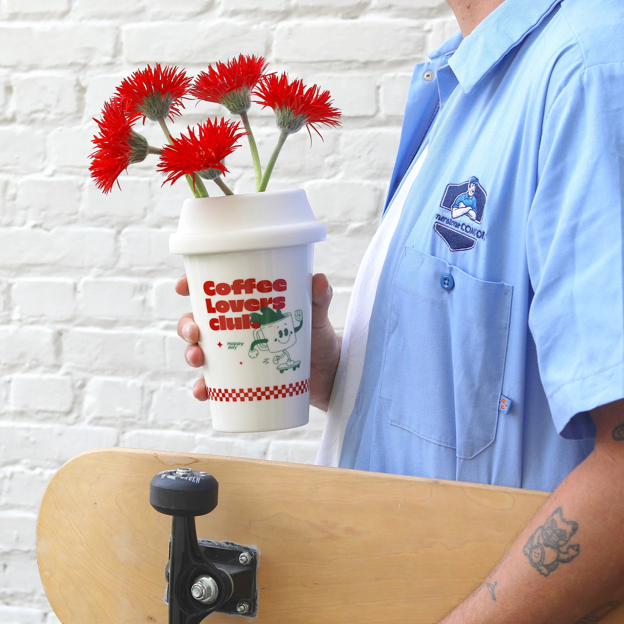 Een persoon in een blauw shirt houdt een skateboard en de Fluid Market Cup of Coffee Vaas vast, in de vorm van een koffiekopje met rode bloemen en de afbeelding "Coffee Lovers Club". Dit Fluid Market decor is perfect voor elke koffieliefhebber, tegen een witte bakstenen muur.