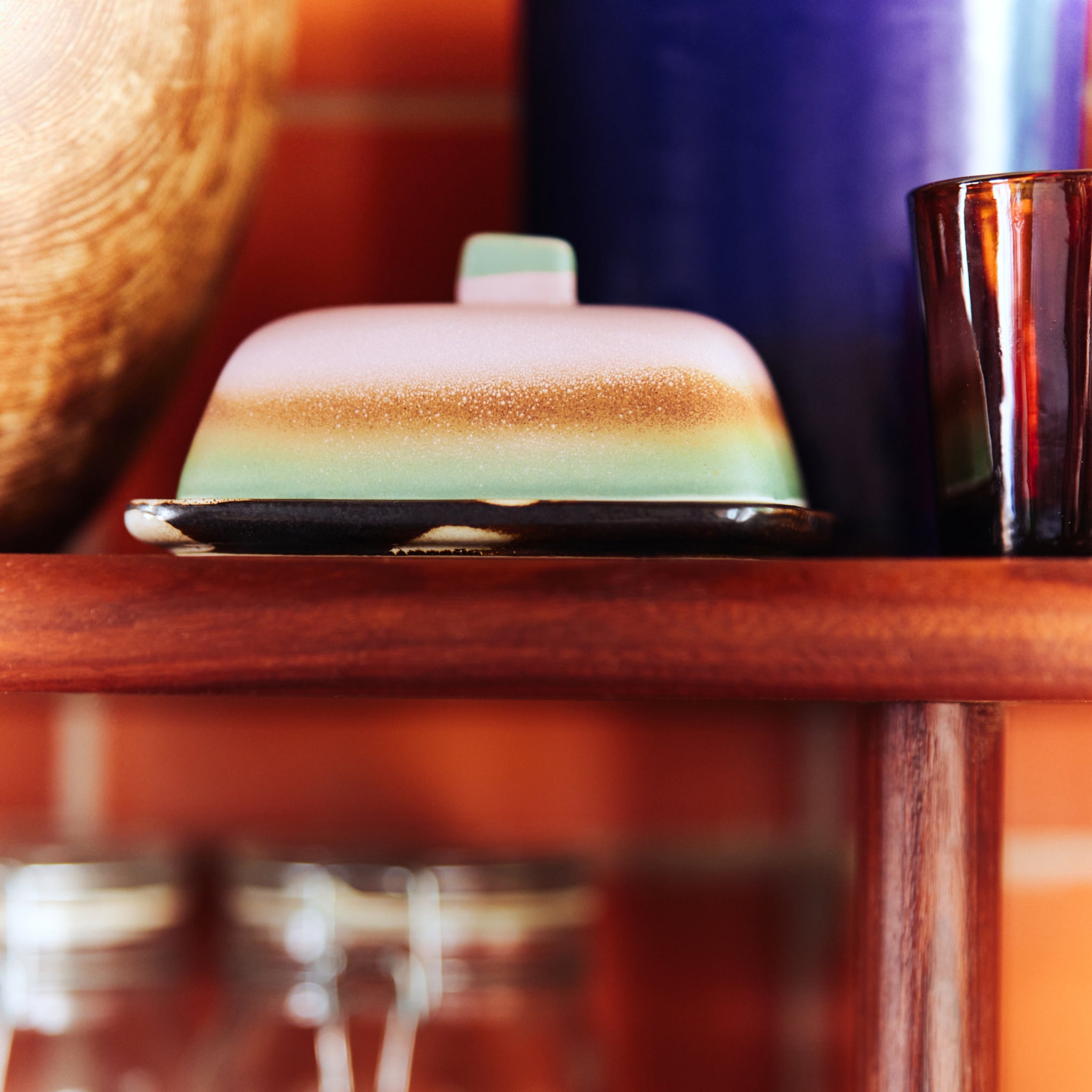 Het HK Living 70s Ceramics Botervlootje Mercury botervlootje staat op een houten plank, met daarachter een grote blauwe vaas en bruin glas, terwijl de glazen potten eronder de retro vibe weerspiegelen tegen een muur van rode bakstenen.