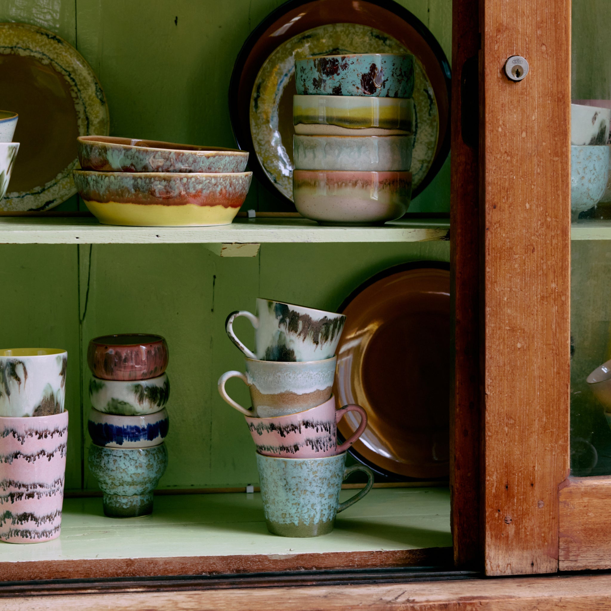 Een lichtgroene houten kast met een glazen deur toont de HK Living 70s Ceramics Cappuccinokop Dada 4 st. set, kommen en borden - allemaal met aardgroene, roze, beige en blauwe glazuren van HK Living.