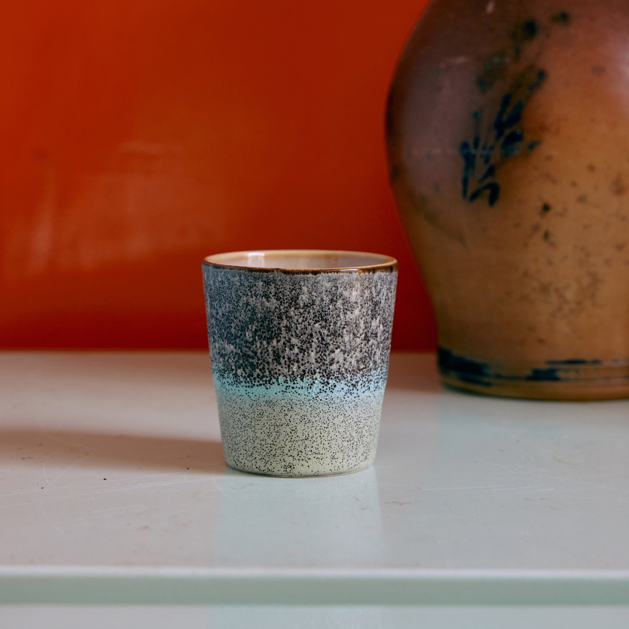 Een HK Living 70s Ceramics Koffiekop Thicket met een gestructureerde, verlopen afwerking van wit naar donkergrijs rust op een bleek oppervlak voor een oranje muur, naast een bruine keramieken vaas versierd met een zachtblauw bloemmotief.