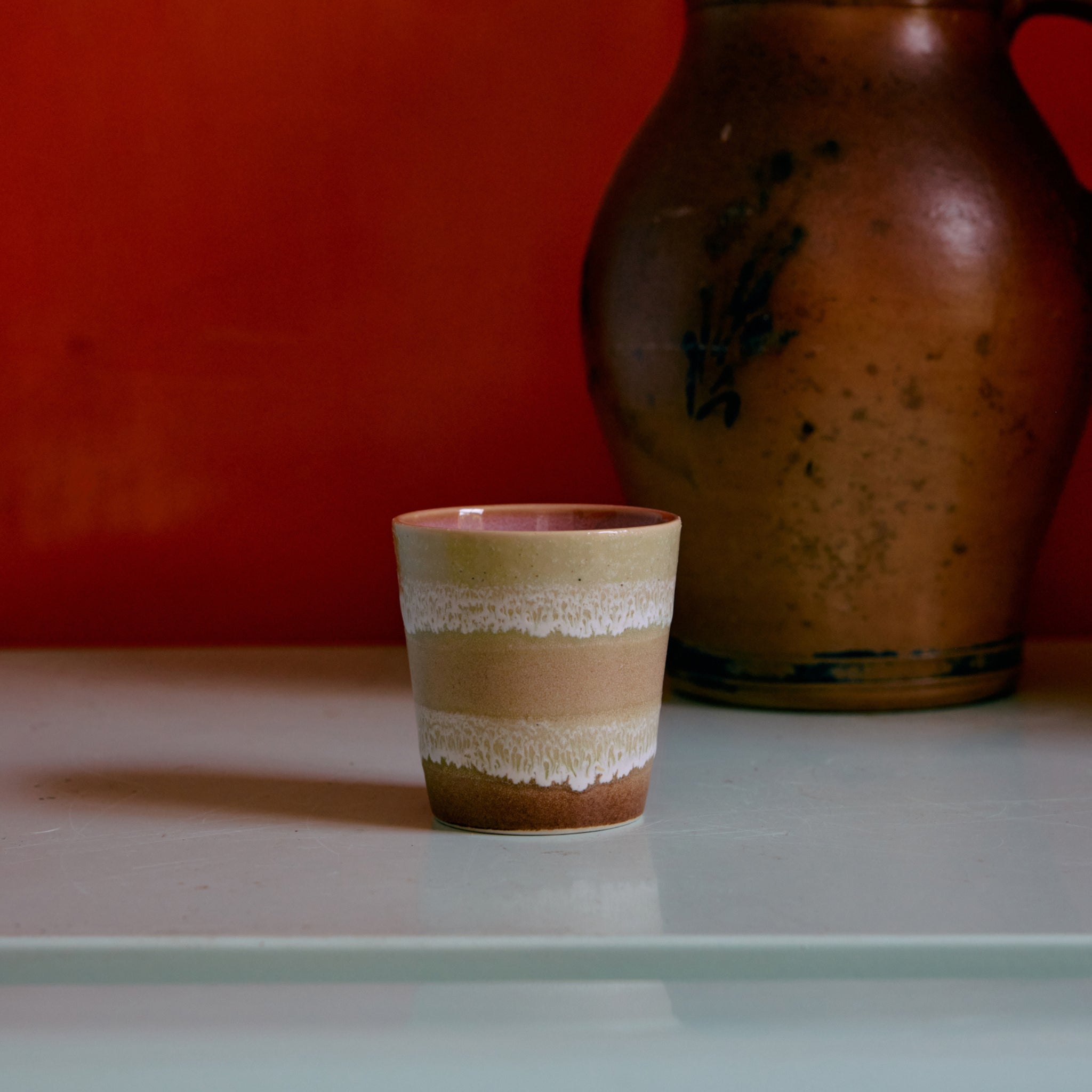 Een kleine HK Living 70s Ceramics Koffiekop Umber, bruin met een crèmekleurig golvend patroon, staat op een licht oppervlak. Daarachter staat een grote bruine aardewerk kan tegen een rood-oranje muur, die de HK Living stijl benadrukt.