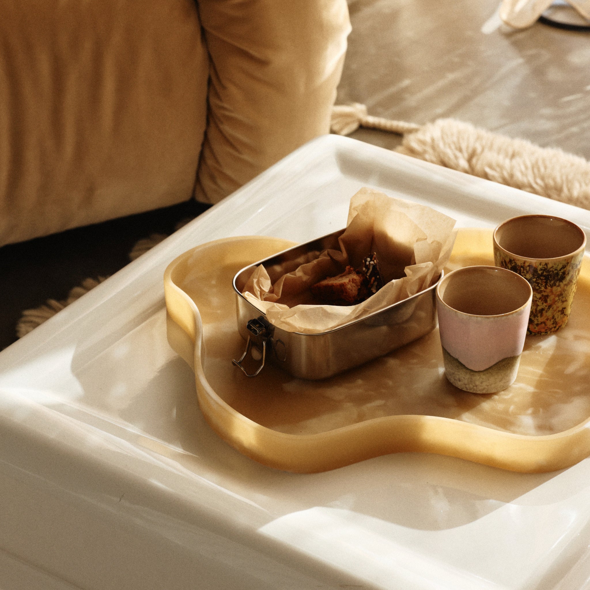 Een gouden dienblad op een witte tafel bevat een metalen bak met in papier gewikkelde etenswaren en twee HK Living 70s Ceramics Koffiekop Bramble mokken. Op de achtergrond verschijnen een beige bank en een donzig vloerkleed in warm natuurlijk licht.