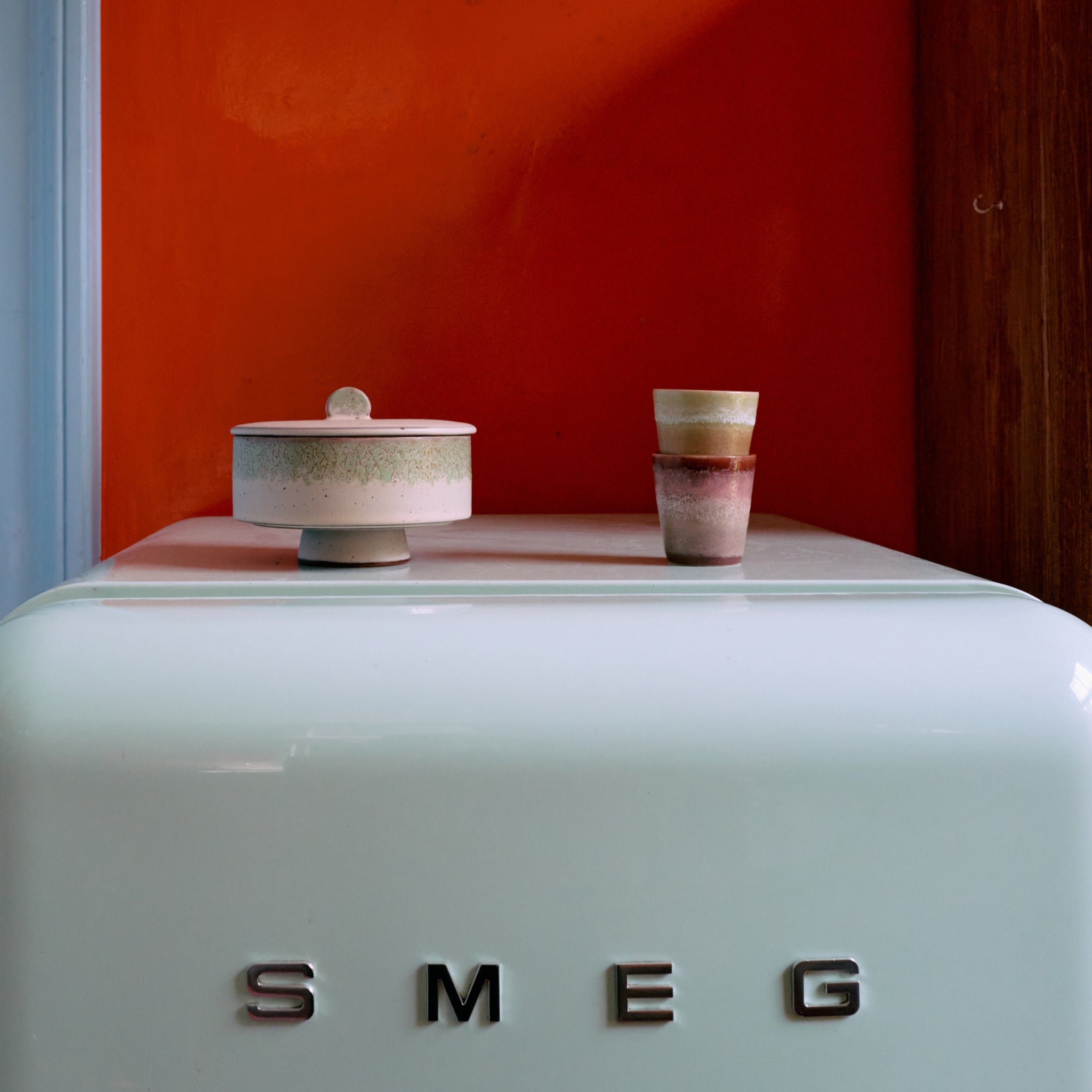 Een witte SMEG koelkast met daarop een keramische pot en twee HK Living 70s Ceramics Koffiekop Umber kopjes, tegen een oranje muur en een houten paneel aan de rechterkant.