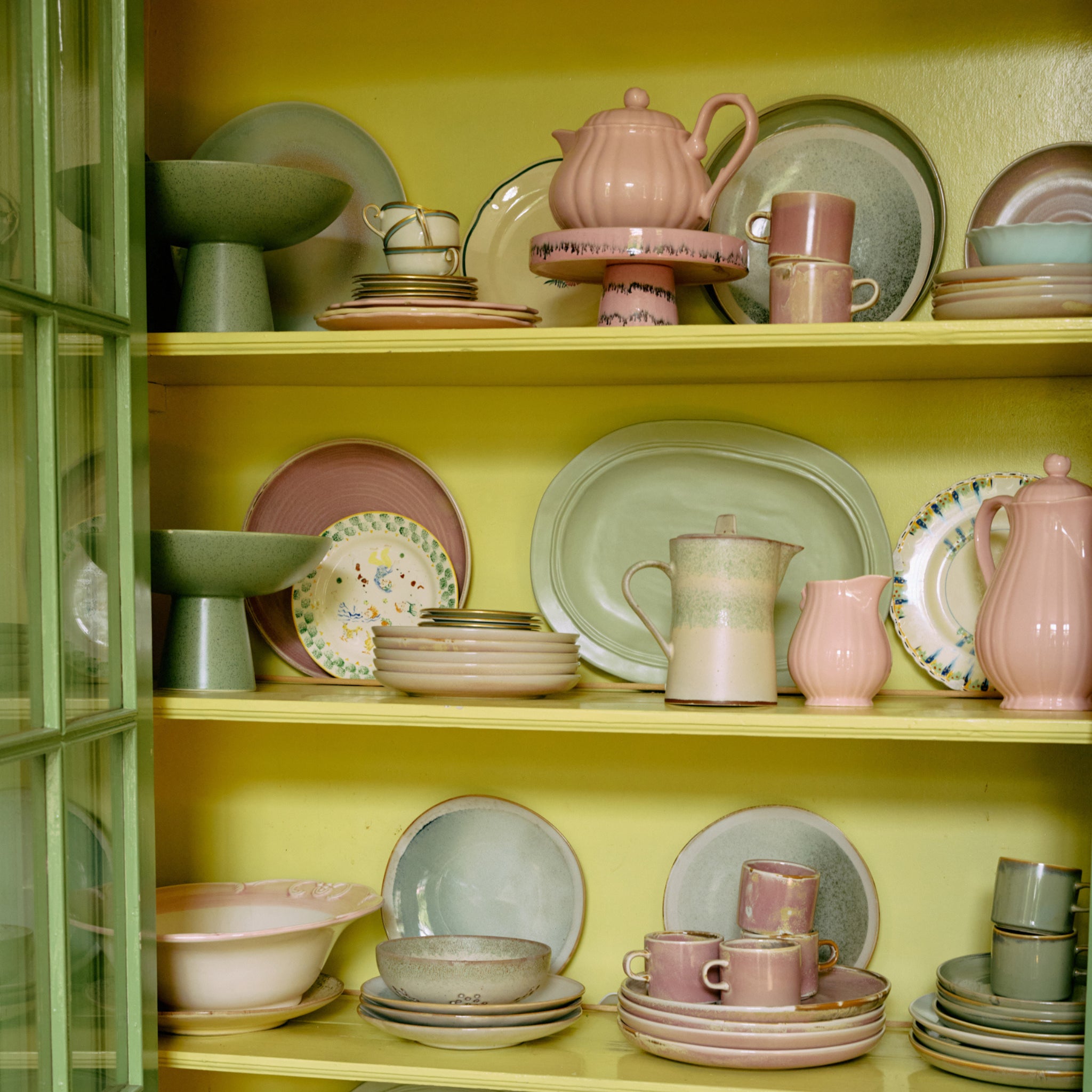 HK Living 70s Ceramics Kom op Voet Bubble, mokken, theepotten en schaal op voet in pastelroze, groen en beige zijn kunstig gerangschikt op planken tegen een gele achtergrond in een glazen deurkast.