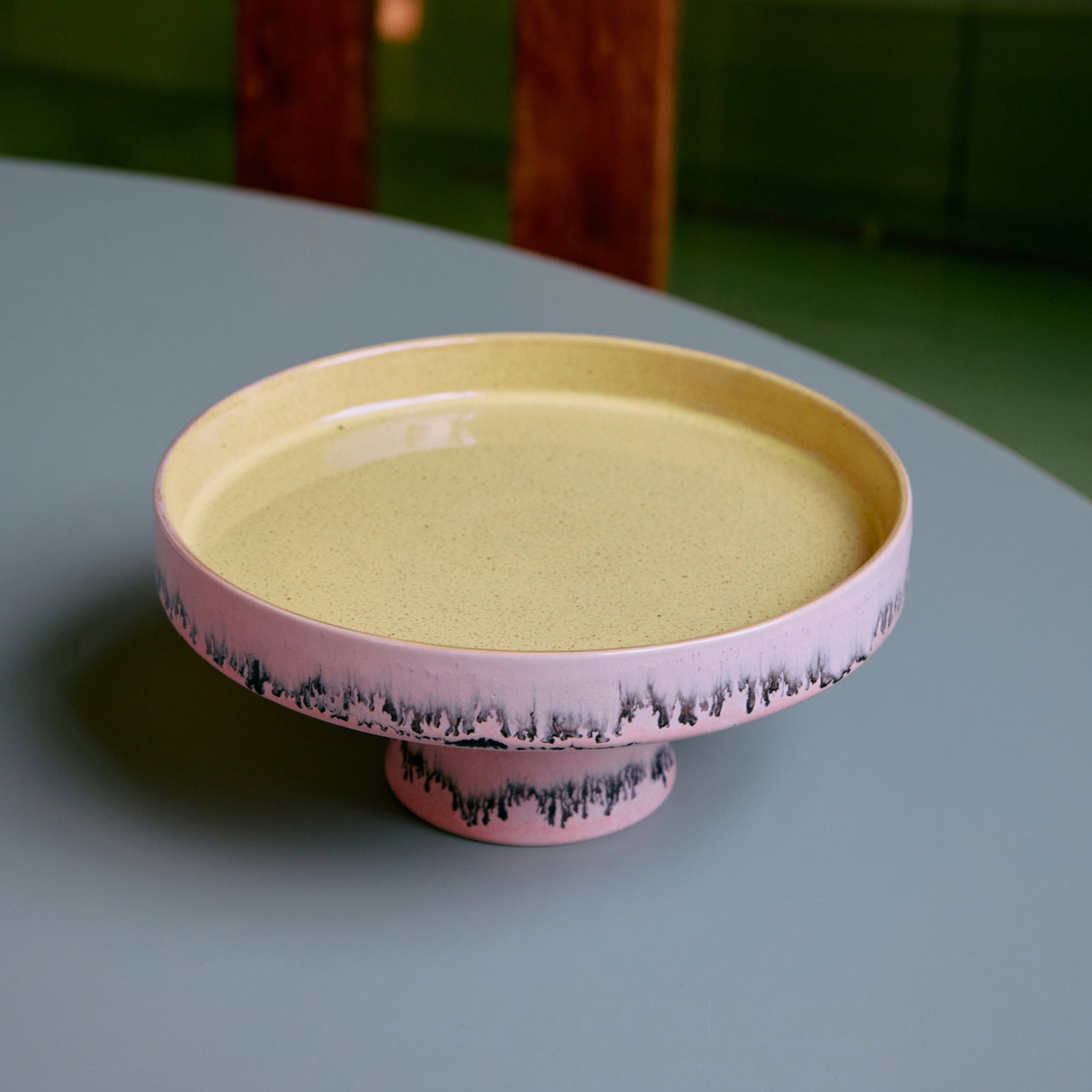 De HK Living 70s Ceramics Kom op Voet Bubble is een keramische taartstandaard op voet met een gele bovenkant, roze basis en donker onregelmatig randpatroon, weergegeven op een lichtblauw oppervlak.