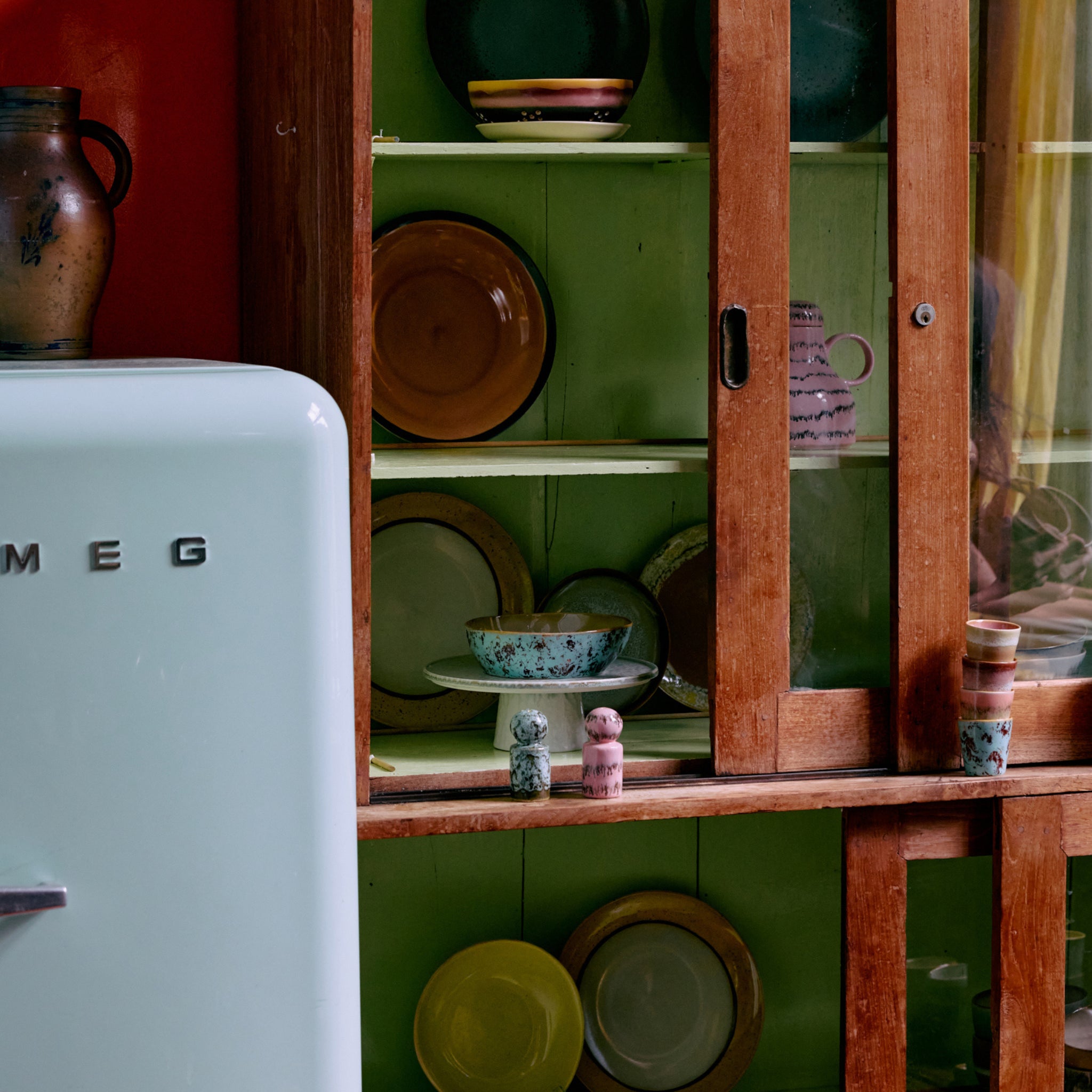 Een pastelgroene SMEG koelkast staat naast een vintage kast vol kleurrijke borden en HK Living 70s Ceramics Theekop Baroque 2 st., waardoor een gezellige retro keuken ontstaat.