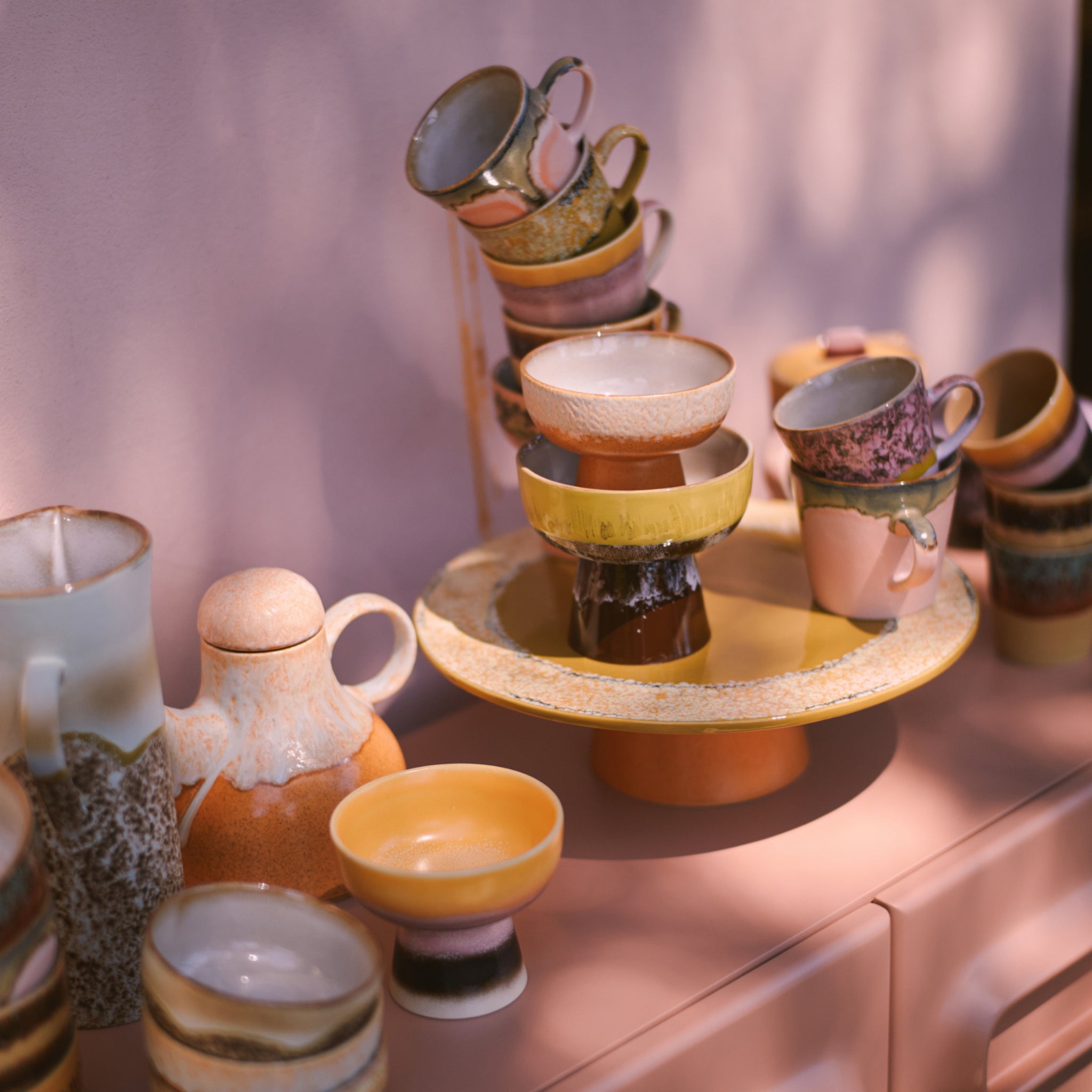 Een levendige set HK Living 70s Ceramics kopjes, kommen en een theepot is gerangschikt op roze, met enkele kopjes gestapeld op de HK Living 70s Ceramics Plateau/Taartschaal Papaya. Zacht natuurlijk licht werpt warme, artistieke schaduwen.