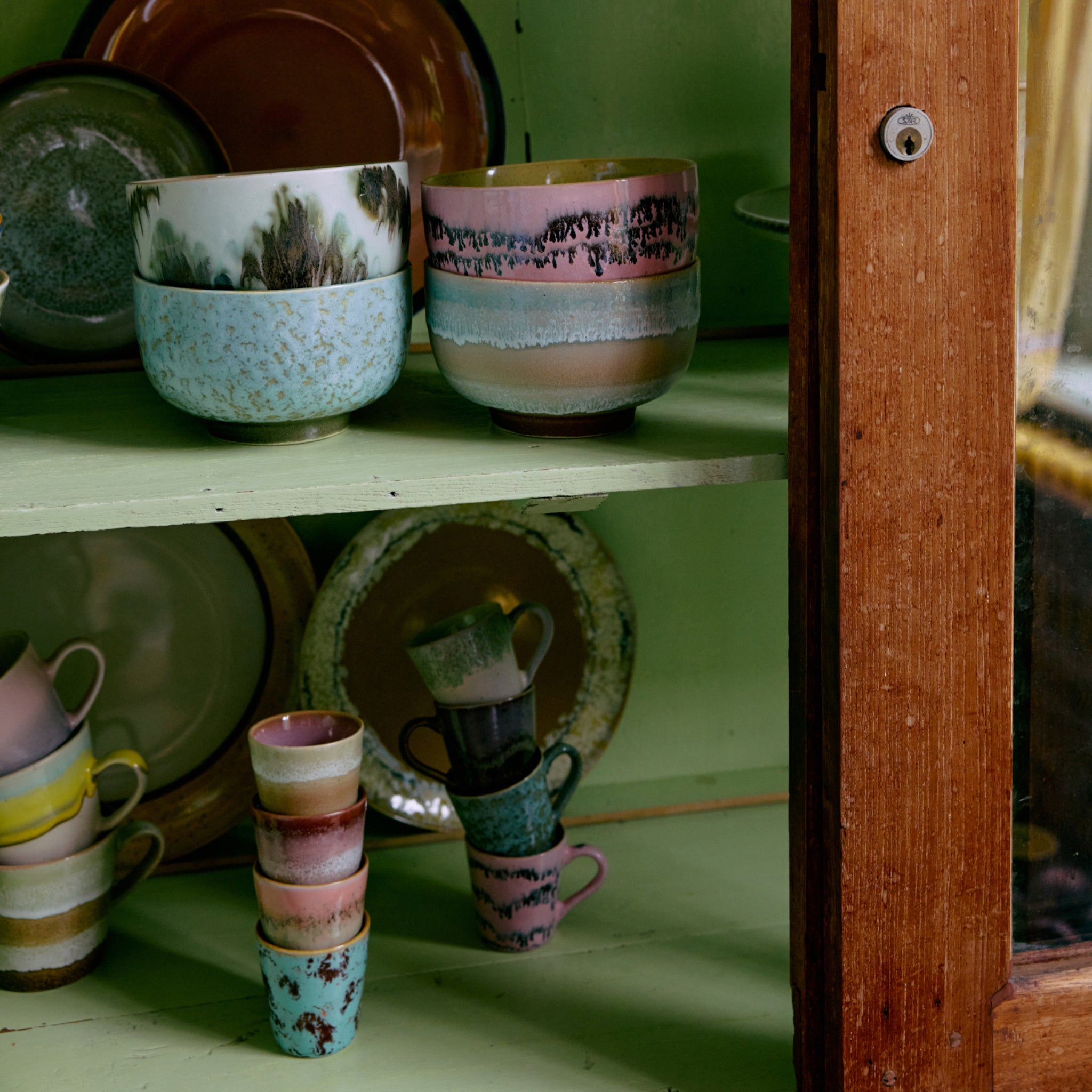 Een houten kast met een glazen deur toont HK Living 70s Ceramics Ristretto Mok Futurism set van 4, netjes gestapeld op groene planken, naast andere kleurrijke kommen, kopjes en borden van het merk HK Living.