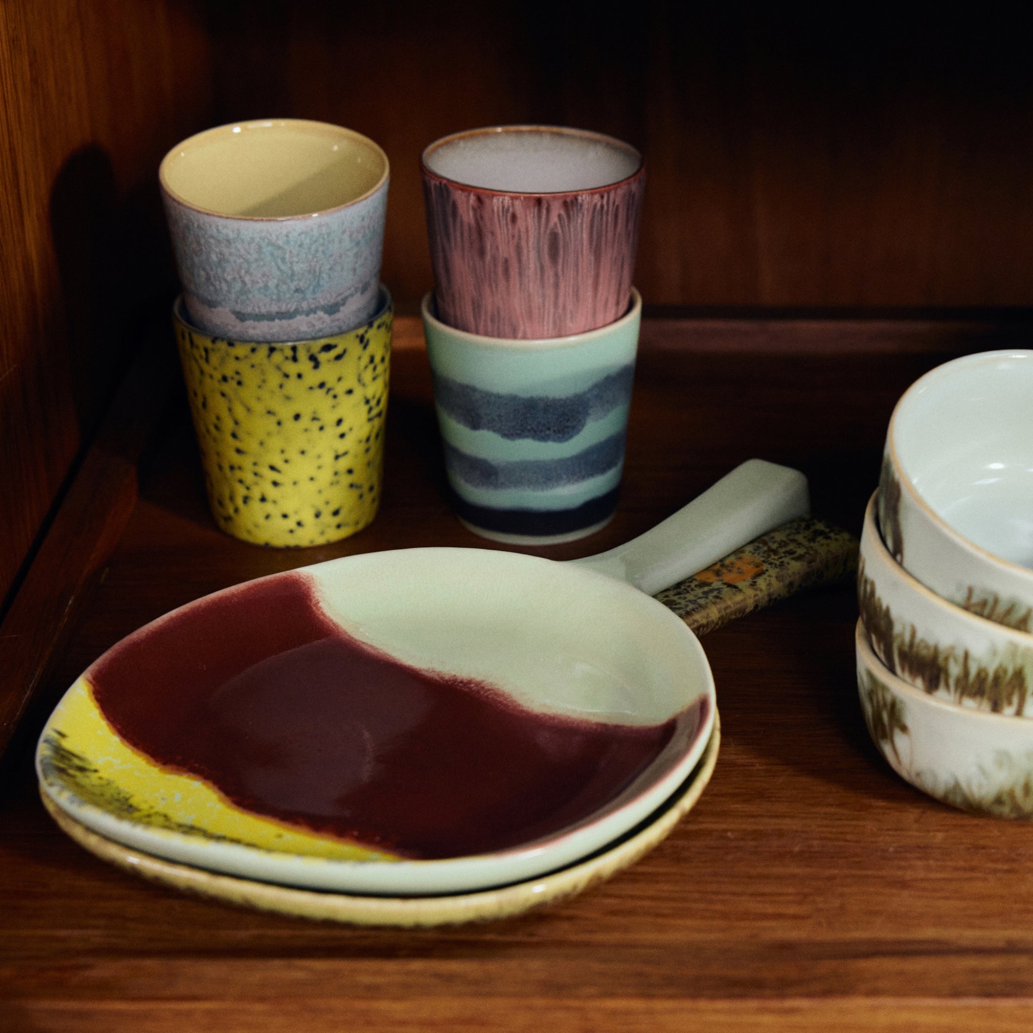 Een levendige reeks HK Living 70s Ceramics Schotel Cherry kopjes en borden staat op een houten plank, met levendige patronen en glazuren geïnspireerd op spikkels, strepen en abstracte kersenmotieven.