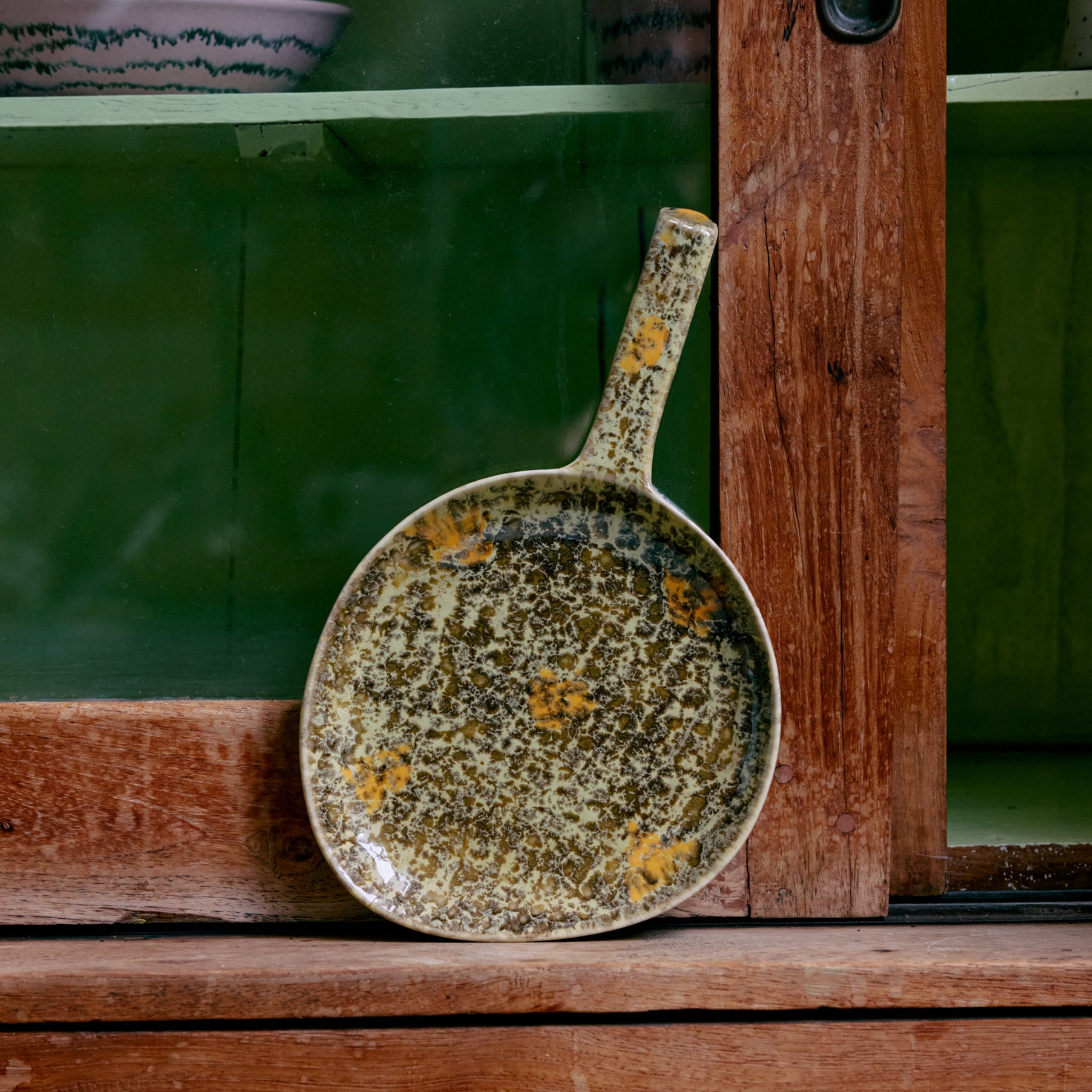 De HK Living 70s Ceramics Schotel Fjord, met een handvat en een gevlekt bruin en geel glazuur, staat rechtop tegen een houten en glazen kast.
