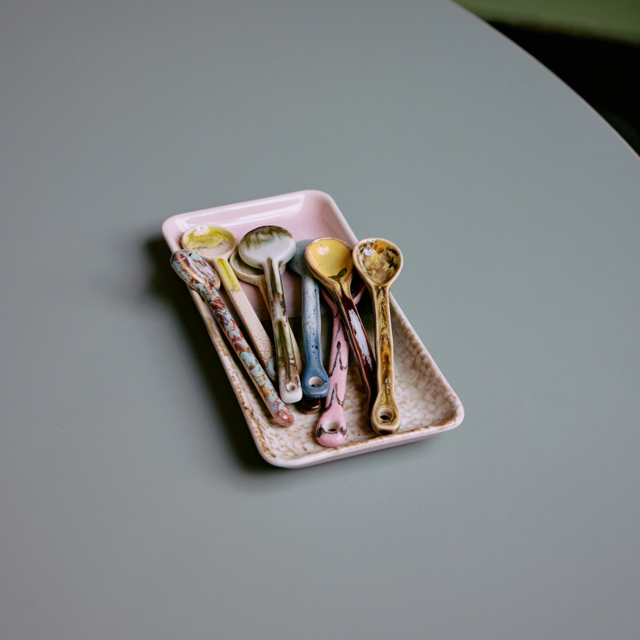 Het HK Living 70s Ceramics Serveerschaaltje Muse bevat verschillende kleurrijke, gespikkelde lepels, allemaal uitgestald op een glad, lichtgrijs oppervlak.