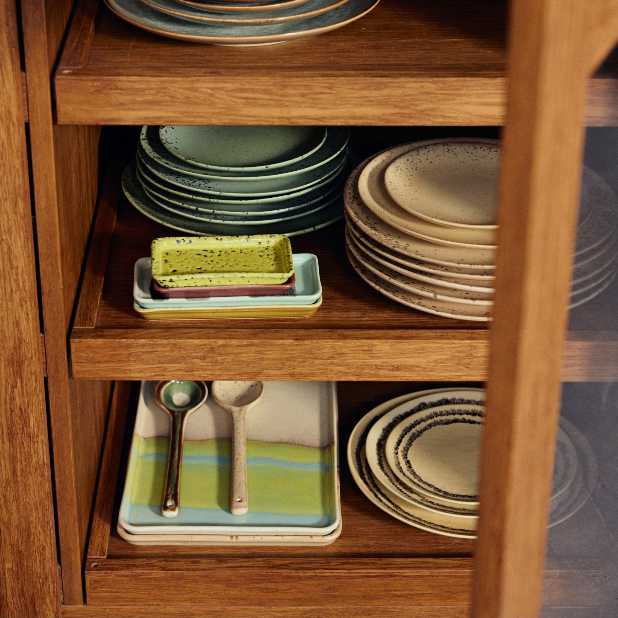 Een houten kast met gestapelde HK Living 70s Ceramics Schaal Glint L, kommen, kleine rechthoekige schaaltjes en twee lepels op een dienblad, allemaal in zachte aardetinten en gedempte kleuren.