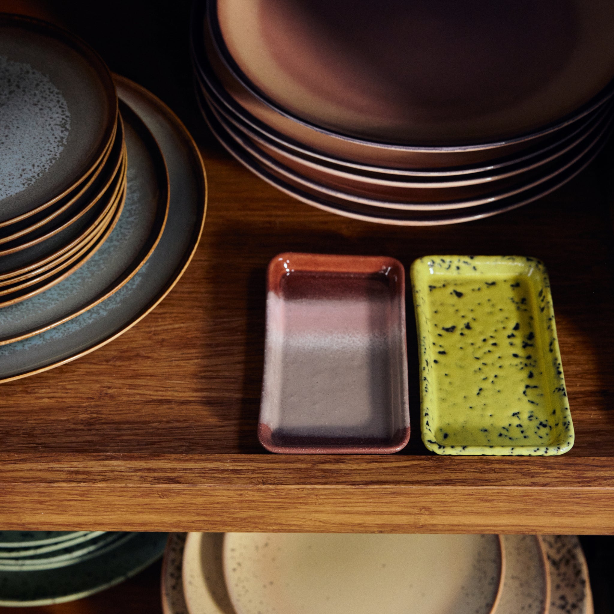 Stapels borden van HK Living keramiek in gedempte tinten staan op houten planken. Op de middelste plank staat een roze bord naast het HK Living 70s Ceramics Serveerschaaltje XS Corn, een geel gespikkeld dienblad.