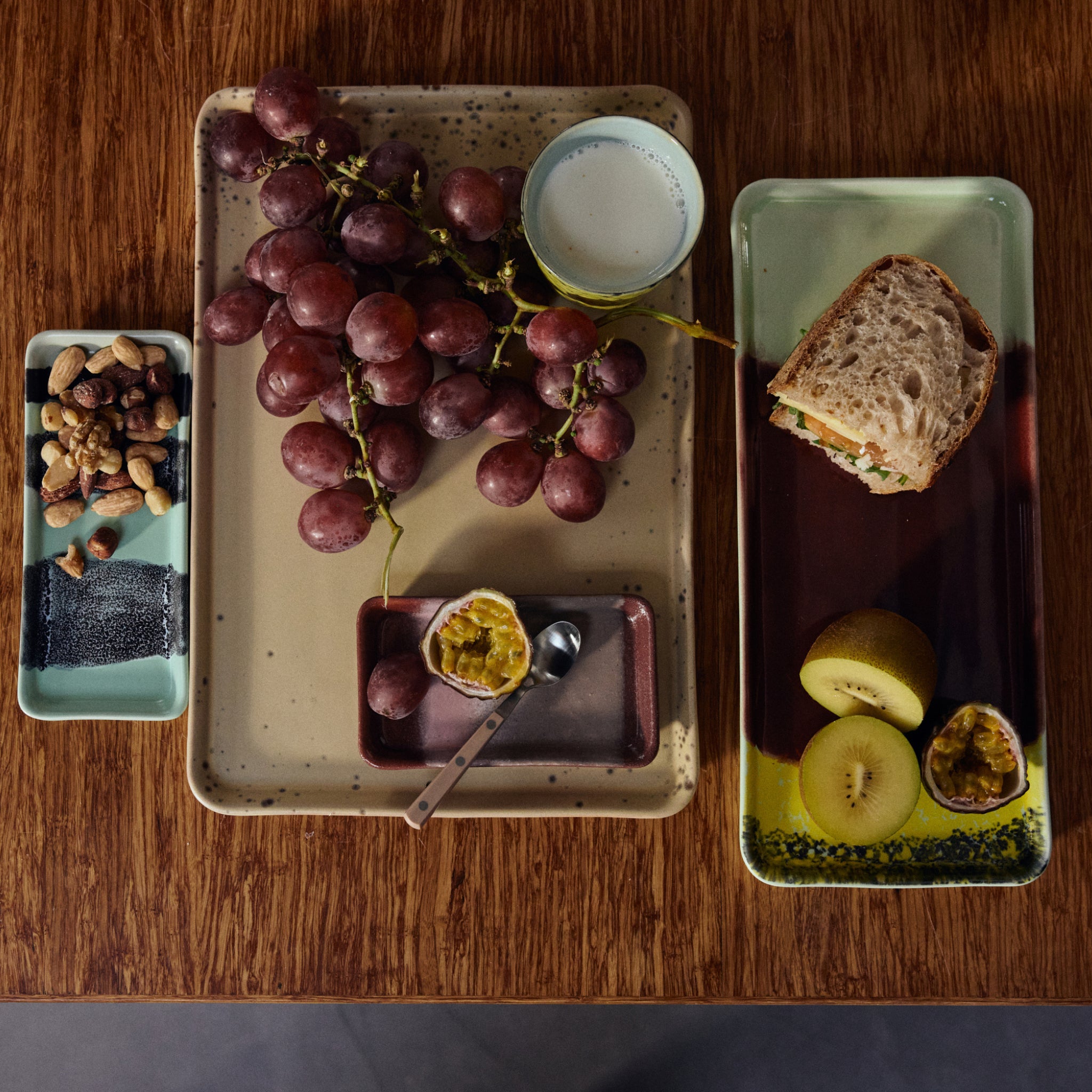 Een houten gedekte tafel met dienbladen met rode druiven, een beker melk, gemengde noten, een boterham, een halve kiwi, kersen, een halve passievrucht met lepel en maanzaadcake op de HK Living 70s Ceramics Cake Schaal Cherry.