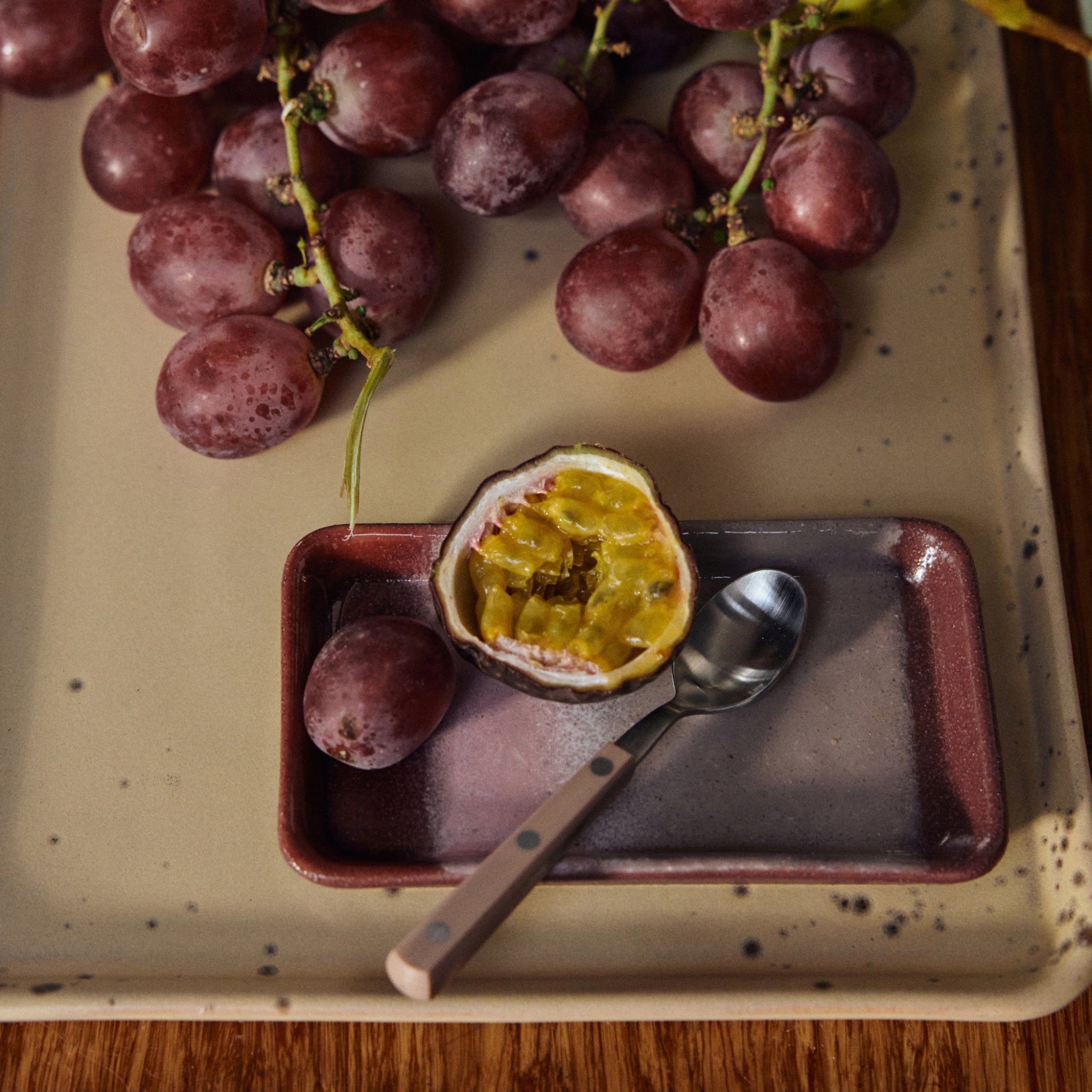 Rode druiven en een halve passievrucht met een lepel zitten op het HK Living 70s Ceramics Serveerschaaltje XS Bloom, geplaatst op een houten ondergrond.