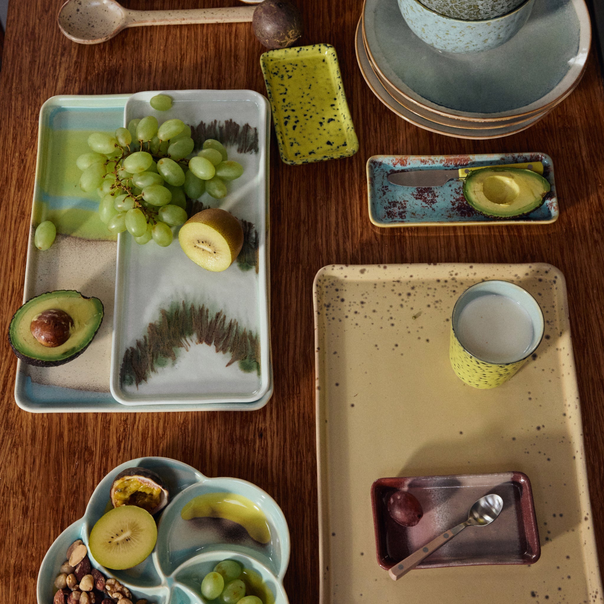 Een houten tafelset met HK Living 70s Ceramics Serveerschaaltje Patina, borden, kopjes en dienbladen bevat groene druiven, avocadohelften, kiwi, gemengde noten en een glas melk. Het HK Living servies heeft aardetinten en pastelkleuren.