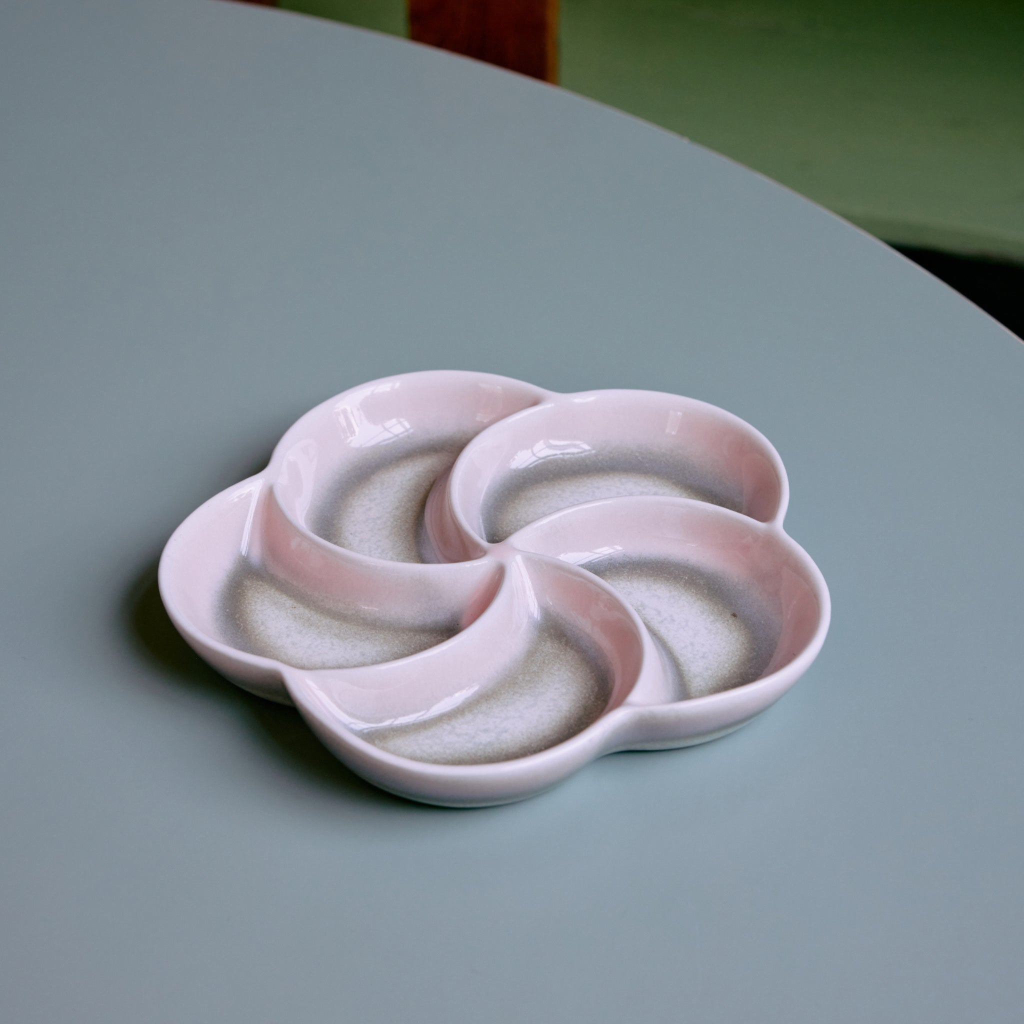 De HK Living 70s Ceramics Snack Schaal Muse is een lichtroze, bloemvormige schaal van keramiek met vier ronde compartimenten, een glanzende afwerking en subtiele spikkels voor retro charme - getoond op een lichtgroen tafelblad.