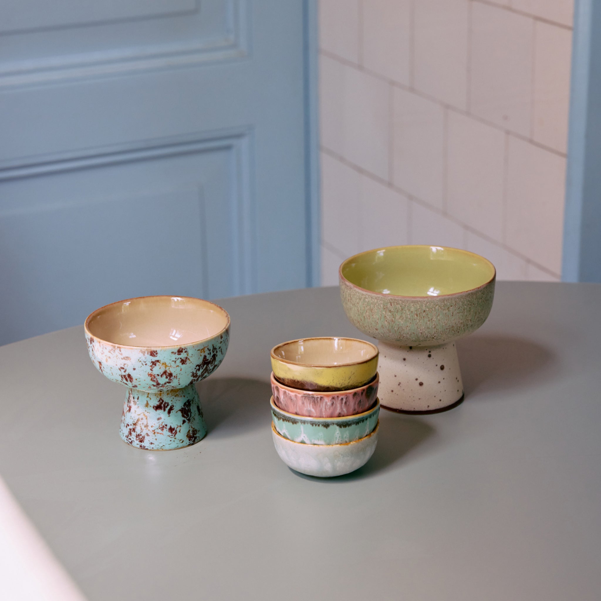 Op een tafel staan twee grote schalen op voet, een daarvan is de HK Living 70s Ceramics Tapas Kom op Voet Slush L met gespikkelde groenachtige patronen, en een stapel van vier kleine kleurrijke kommen, tegen een lichtblauwe muur en betegelde achtergrond.