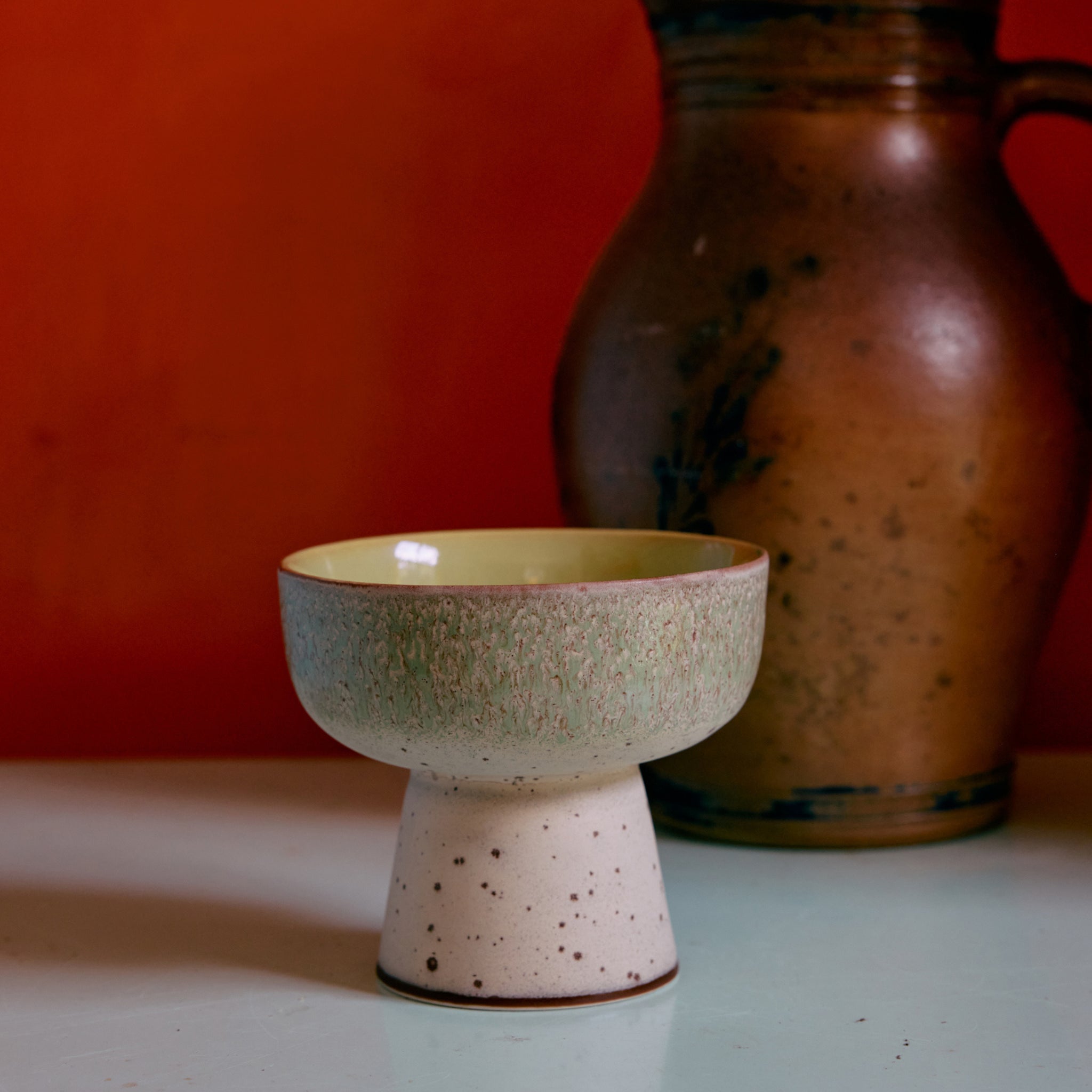 De HK Living 70s Ceramics Tapas Kom op Voet Slush L, met een gespikkelde gebroken witte basis en een kom met groene textuur, staat op een licht oppervlak. Daarachter staat een rustieke bruine kan tegen een oranje muur.