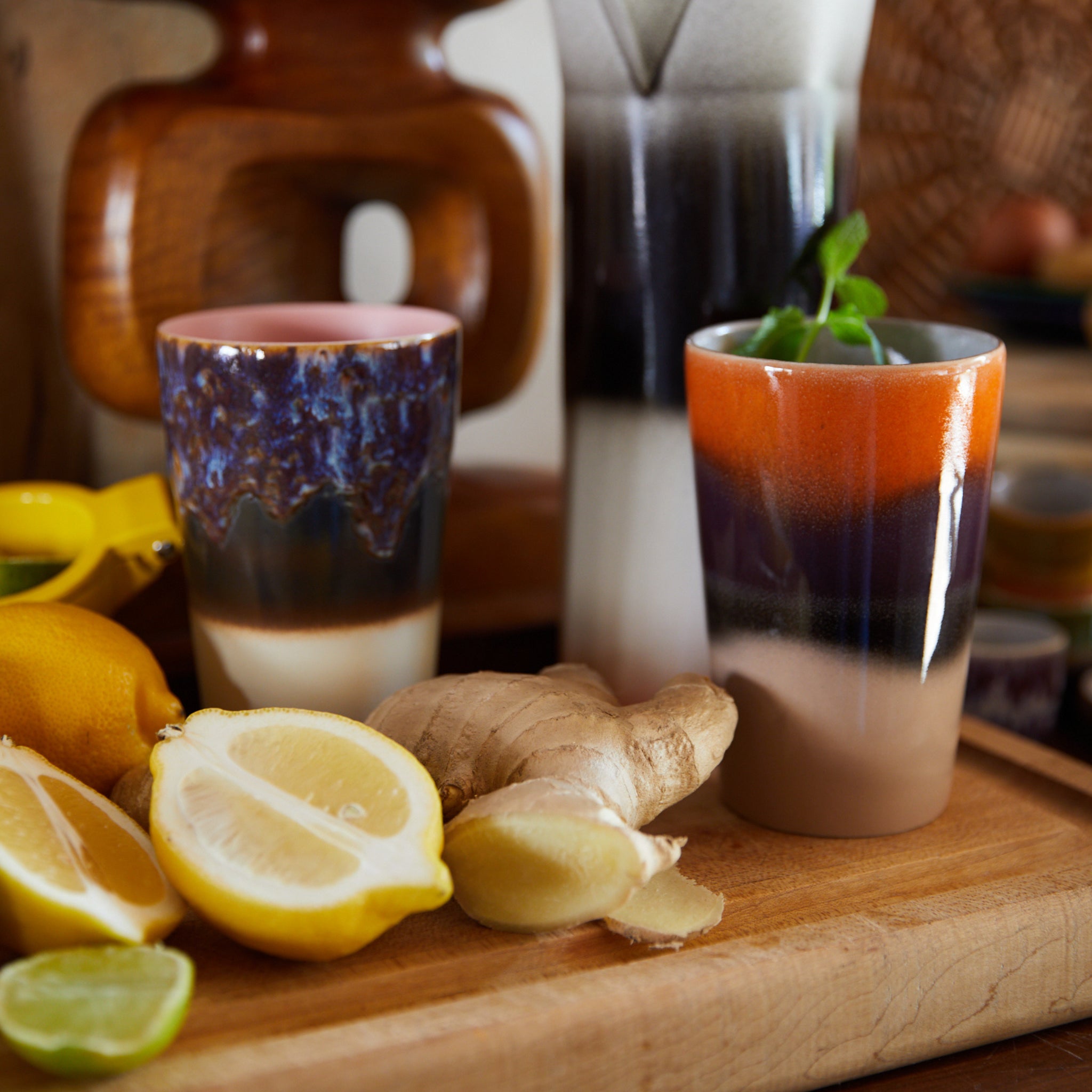 Twee HK Living 70s Ceramics Theekop Dusk mokken staan op een houten plank met gesneden citroen, gember en limoen. Eén mok houdt een munttakje vast, met op de achtergrond keukengereedschap en meer HK Living keramiek.