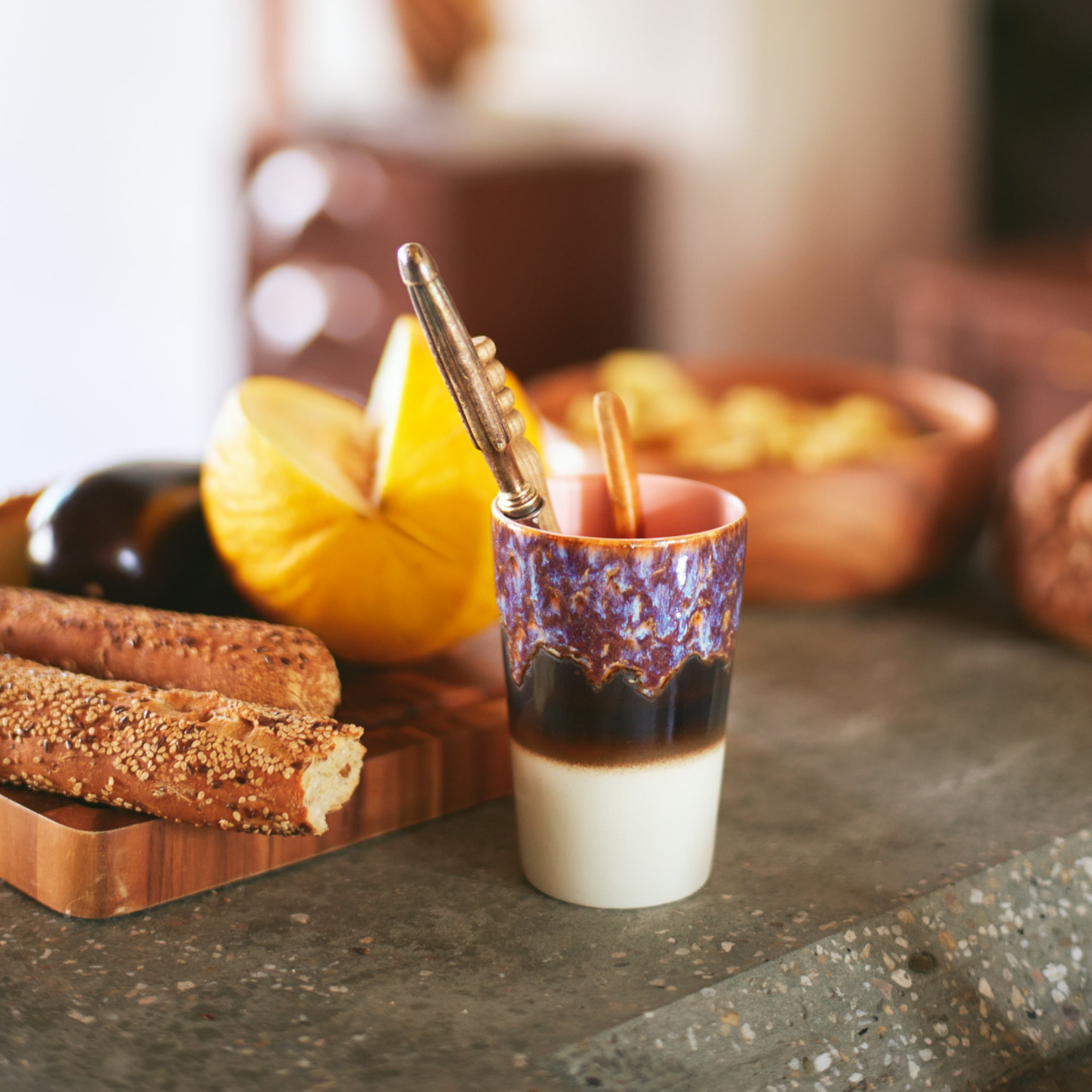 Een HK Living 70s Ceramics Theekop Dusk 2 st. staat op het aanrecht naast broodstengels met zaadjes, een houten dienblad met meloen en een donkere groente, waardoor een gezellige en uitnodigende ontbijtvibe ontstaat.