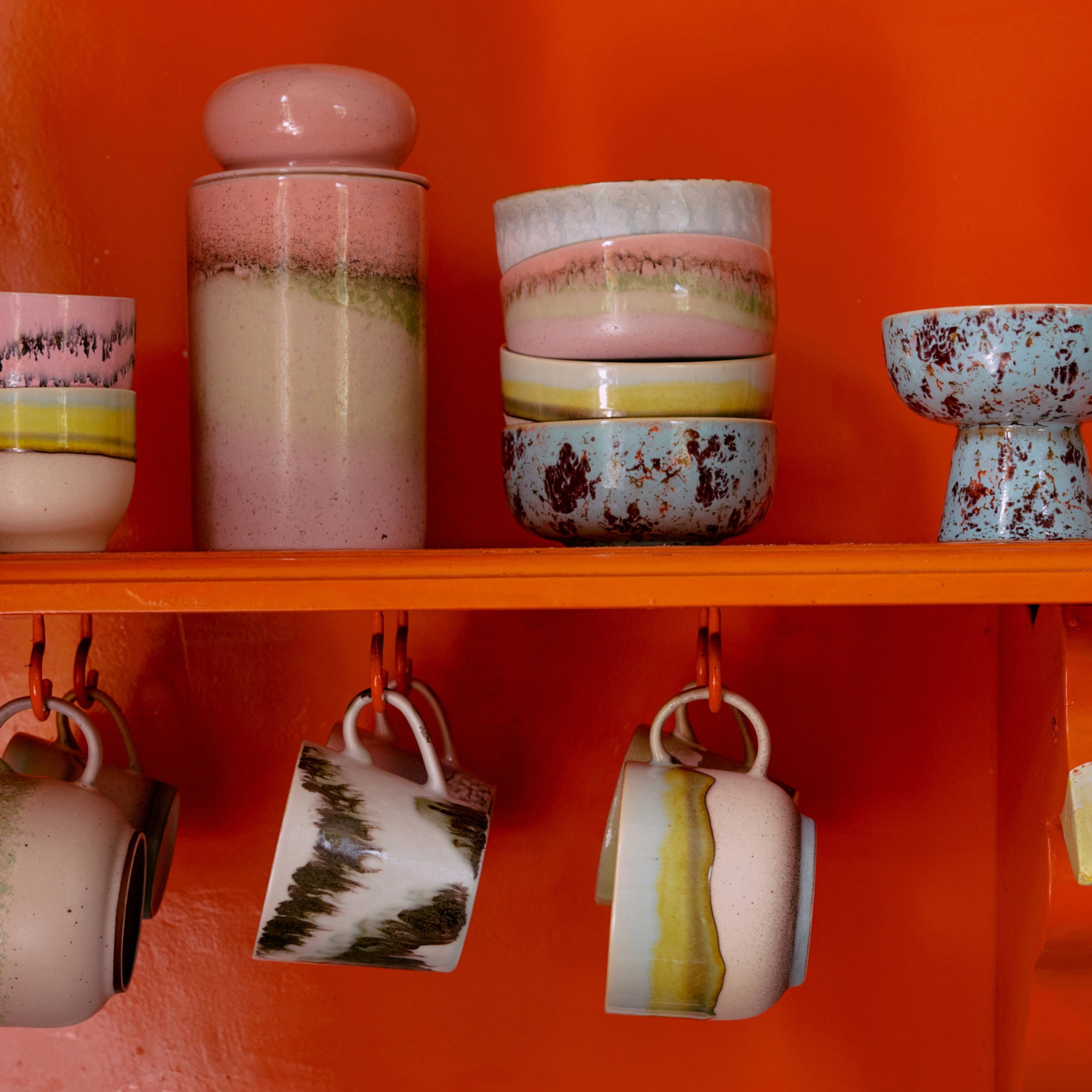Een set HK Living 70s Ceramics, waaronder de Tapas Kom op Voet Patina S, staat op een oranje plank met vier verschillende mokken eronder en gestapelde kommen en een pot erbovenop.