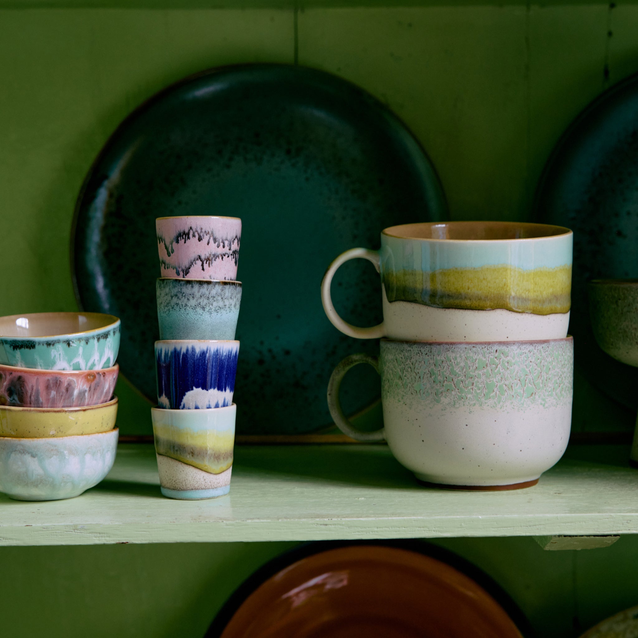 Op een plank staan HK Living 70s Ceramics XS Kom Modernist 4 st., gestapelde kopjes, modernistische kommen met patronen en twee grote mokken - allemaal gerangschikt voor een groene achtergrond met decoratieve borden.
