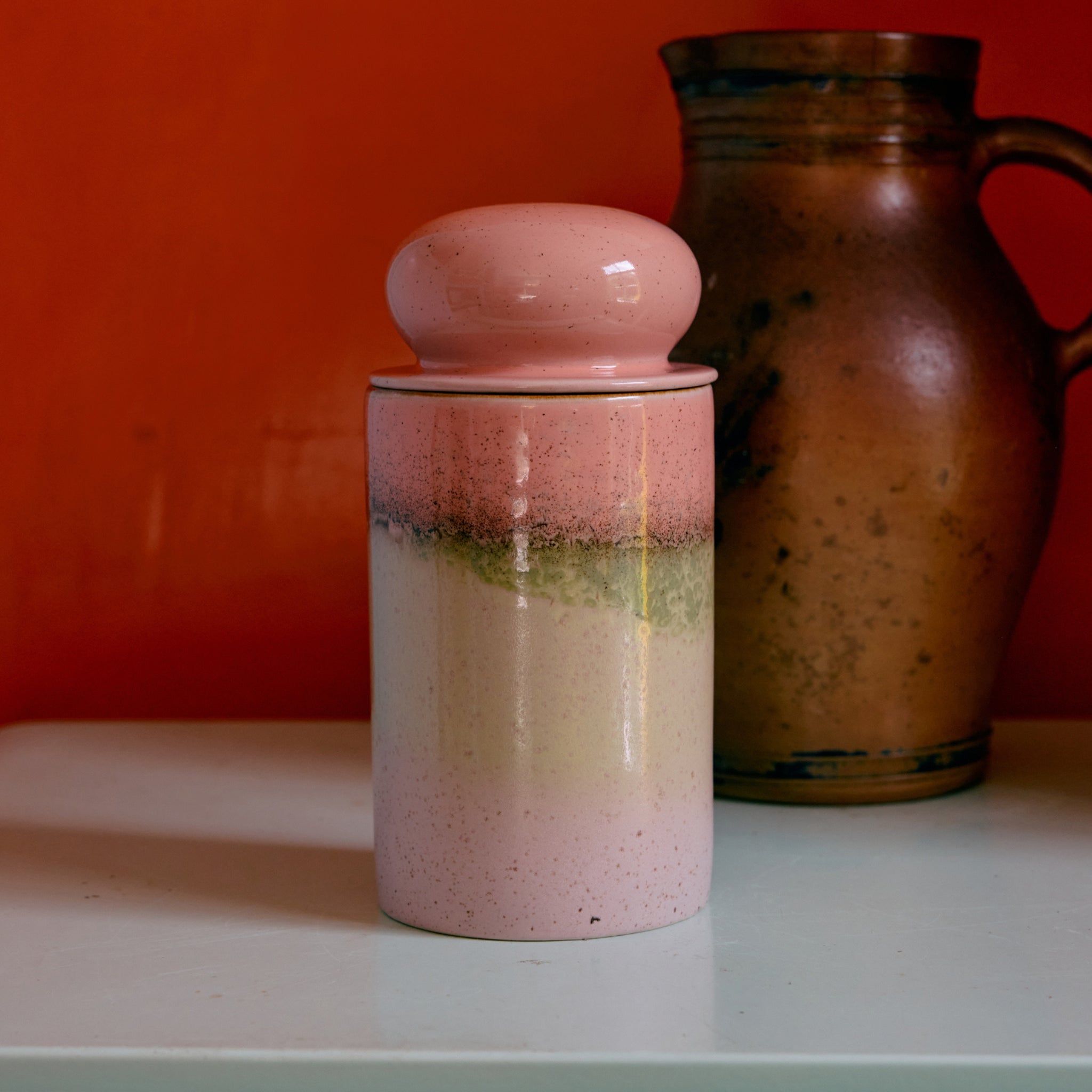 De HK Living 70s Ceramics Voorraadpot Glazuur in roze, met een ronde deksel, rust op een licht oppervlak met een bruine kan erachter en een oranje muur op de achtergrond.