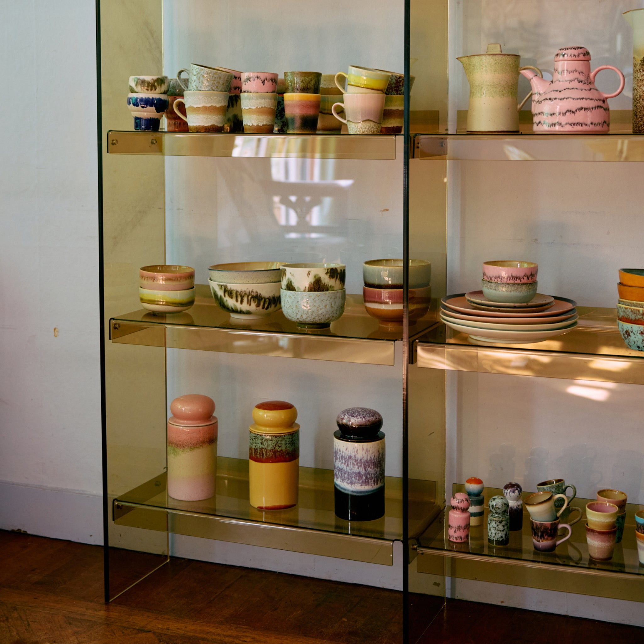 Keramische kopjes, kommen, borden en potten, waaronder de HK Living 70s Ceramics Voorraadpot Glazuur, hebben kleurrijke pastelkleuren en aardse patronen, uitgestald op glazen planken in een goed verlichte kamer met houten vloeren.