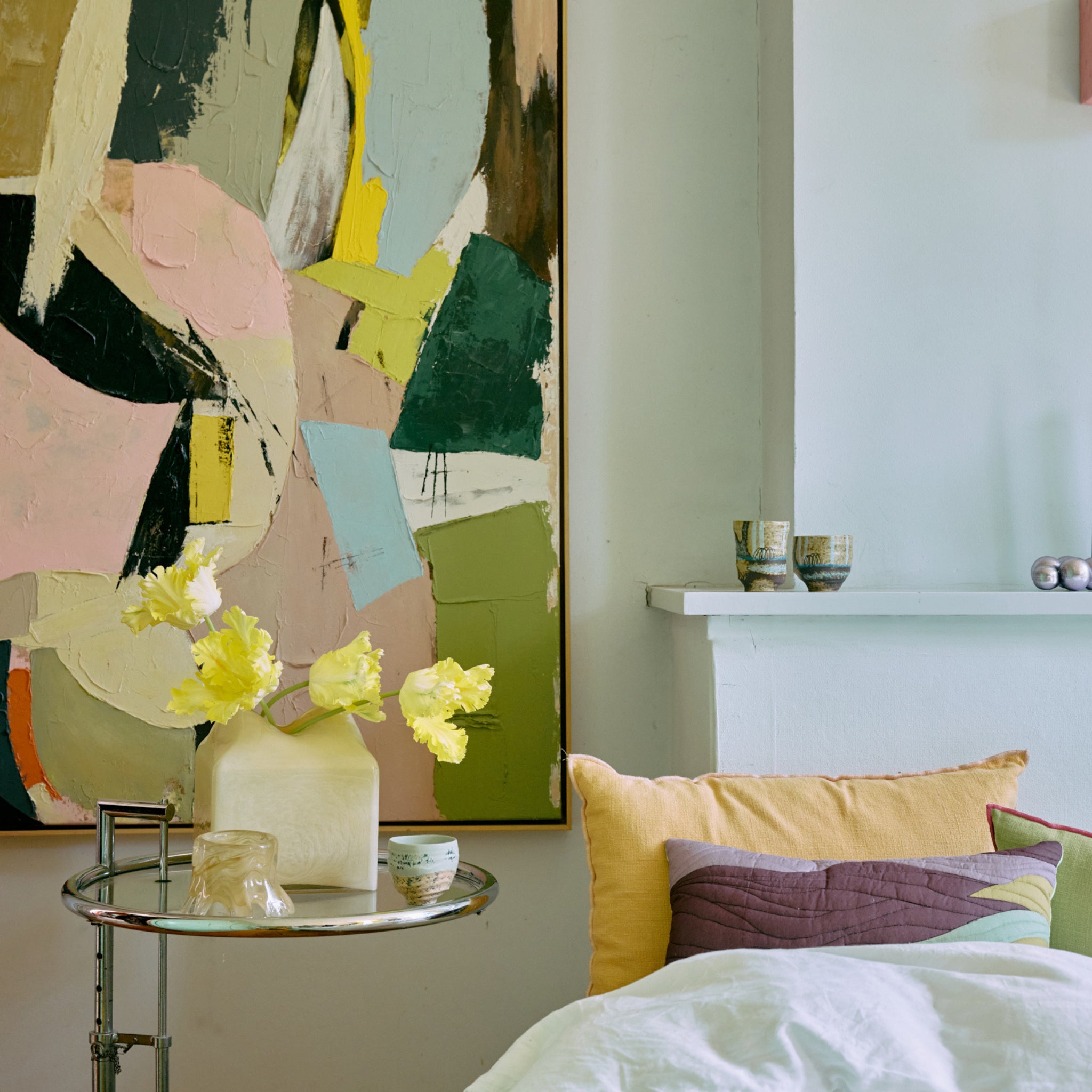 Een moderne slaapkamerhoek met een kleurrijk abstract schilderij, een kleine ronde tafel met een witte vaas met gele bloemen, een HK Living Artist Ceramics Koffiekop 4 st. en pastelkleurige kussens op het bed naast een witte schouw.
