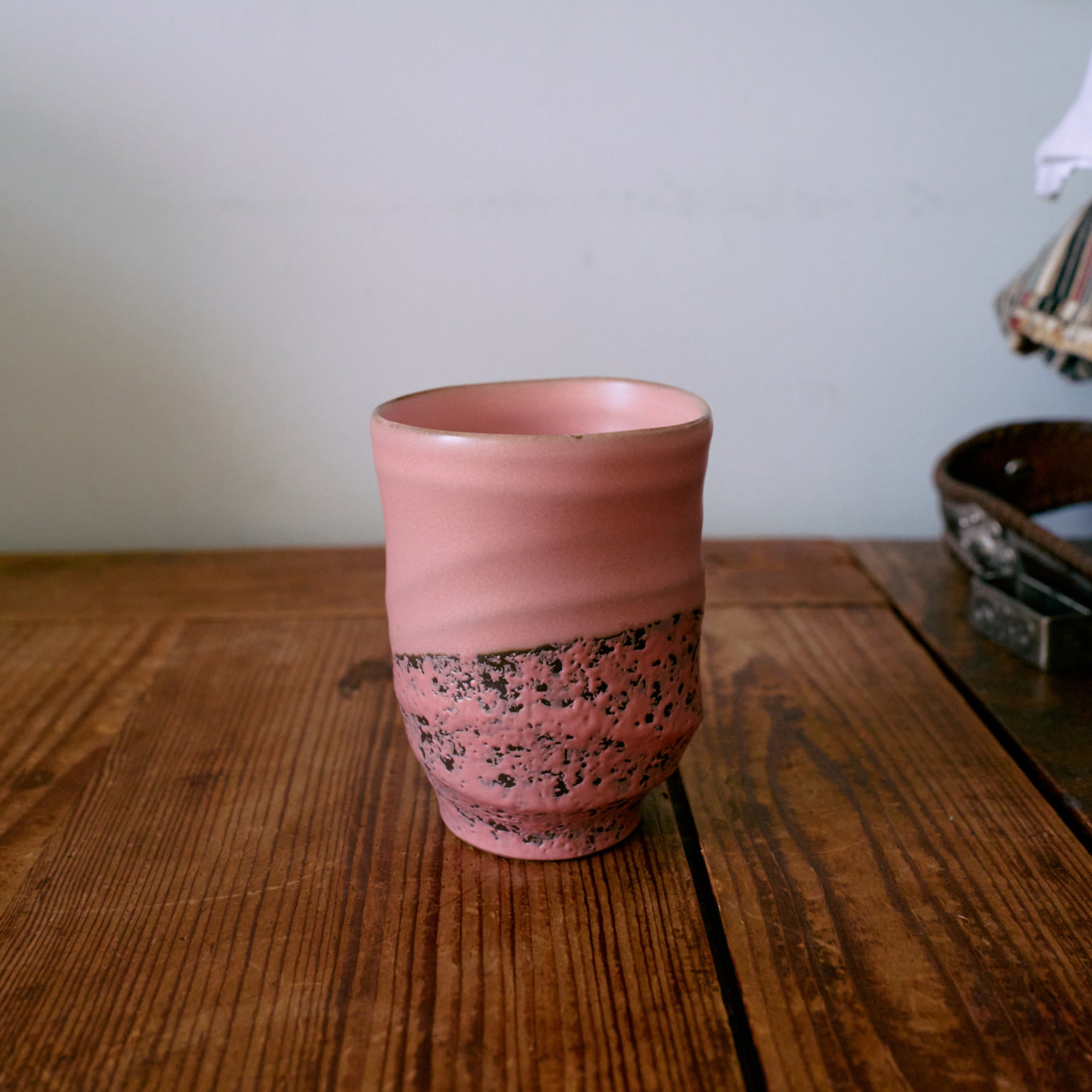 Een HK Living Artist Ceramics Koffiemok uit de 4-delige set, met een roze gestructureerde, gespikkelde onderkant, staat op een houten tafel tegen een lichte muur, met vage objecten op de achtergrond rechts.