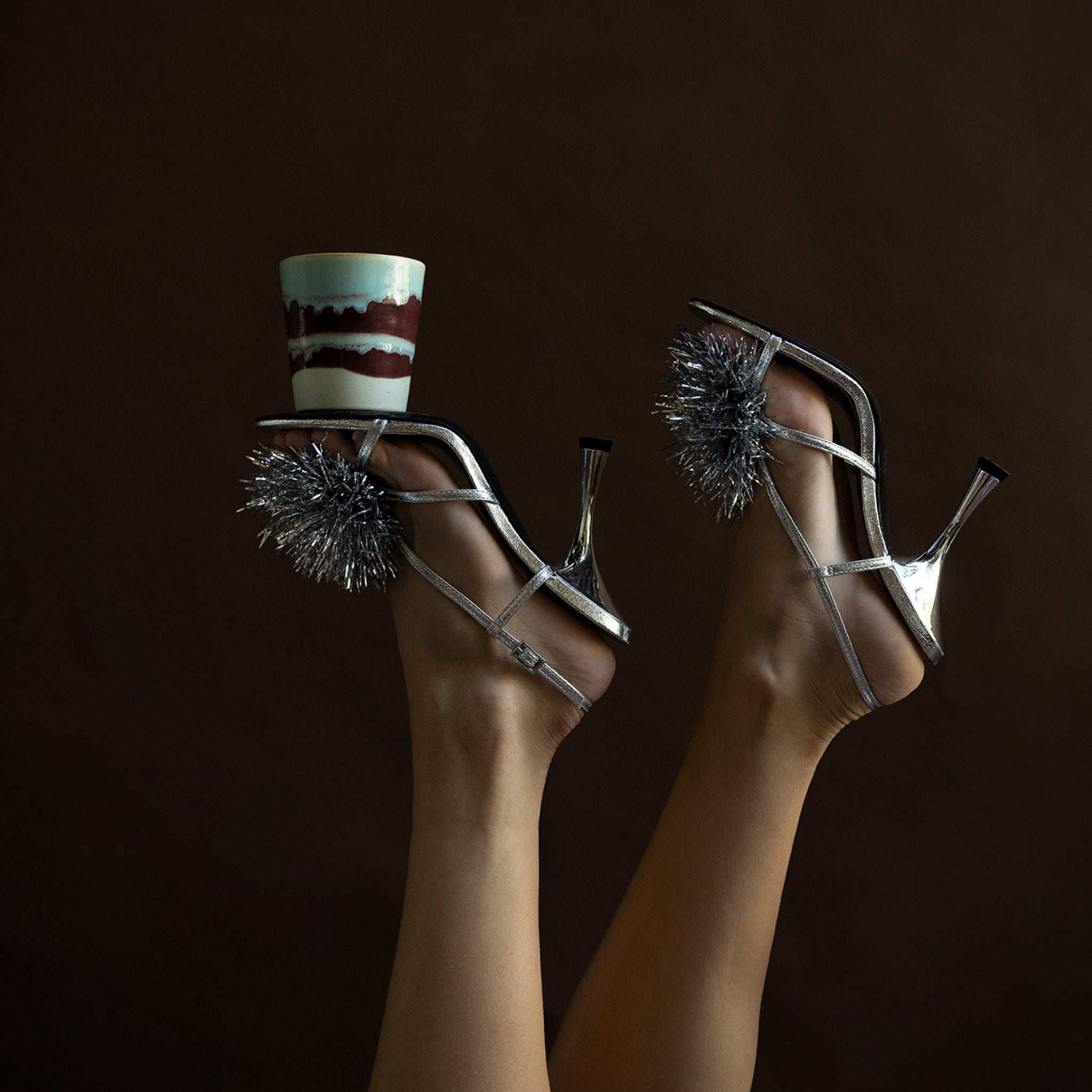 Twee voeten in zilveren sandalen met hoge hakken en metallic pompons steken af tegen een donkere achtergrond. Eén hak balanceert een HK Living Christmas Special Geurkaars Frosted Leaves kaars.