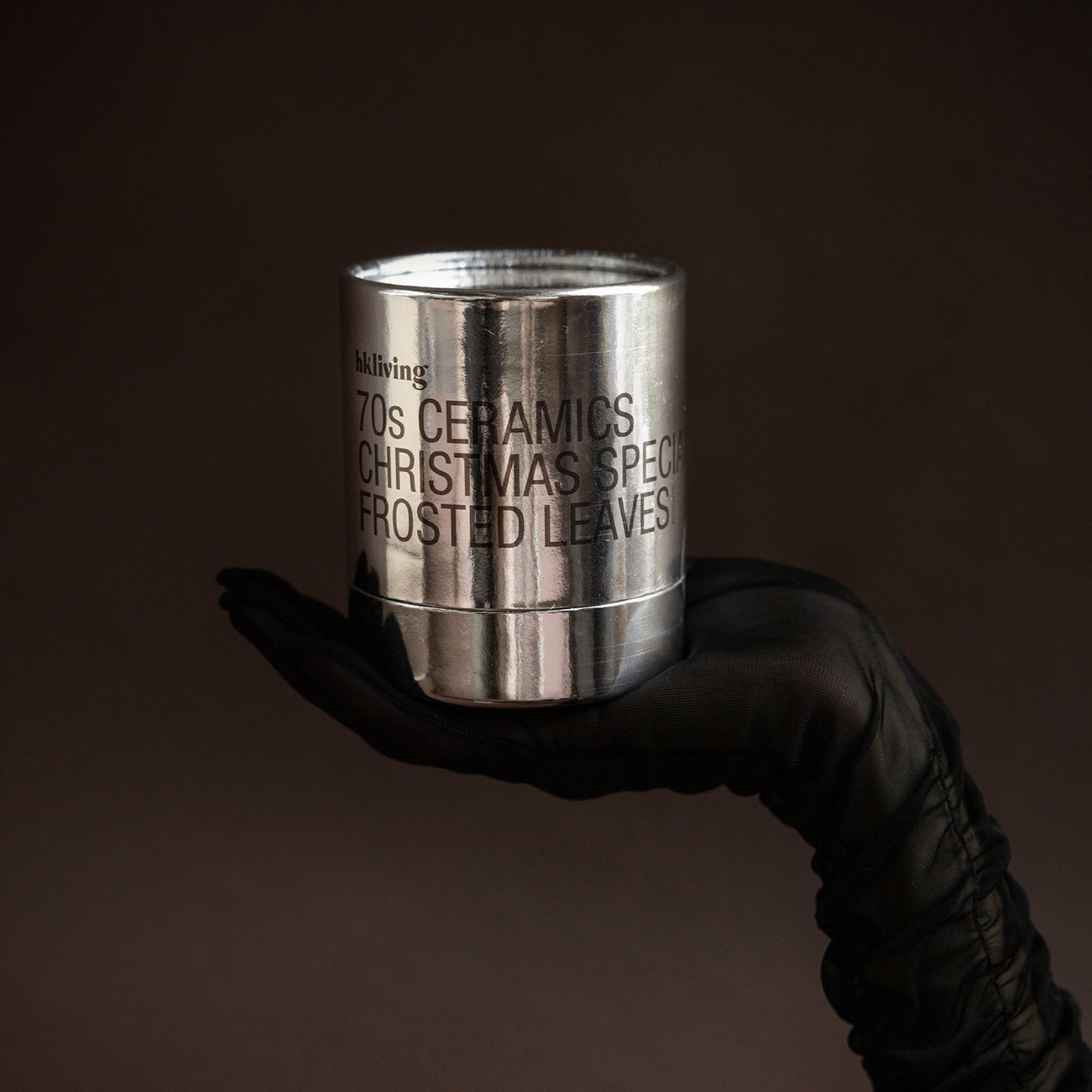 Een gehandschoende hand houdt de HK Living Kerstspecial Geurkaars Frosted Leaves kaarsenpot tegen een donkerbruine achtergrond.