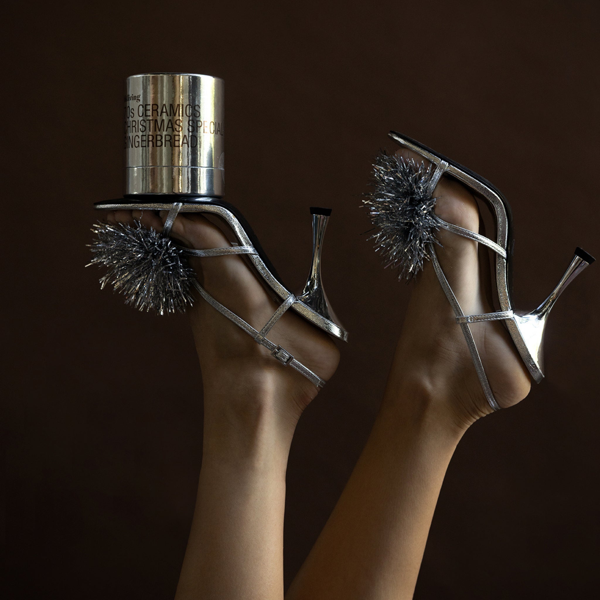 Twee voeten, met glanzend zilveren sandalen met hoge hakken en glinsterende pompons van klatergoud, worden in de lucht geheven. Op één schoen staat een HK Living Christmas Special Geurkaars Peperkoekkaars. De achtergrond is donkerbruin.