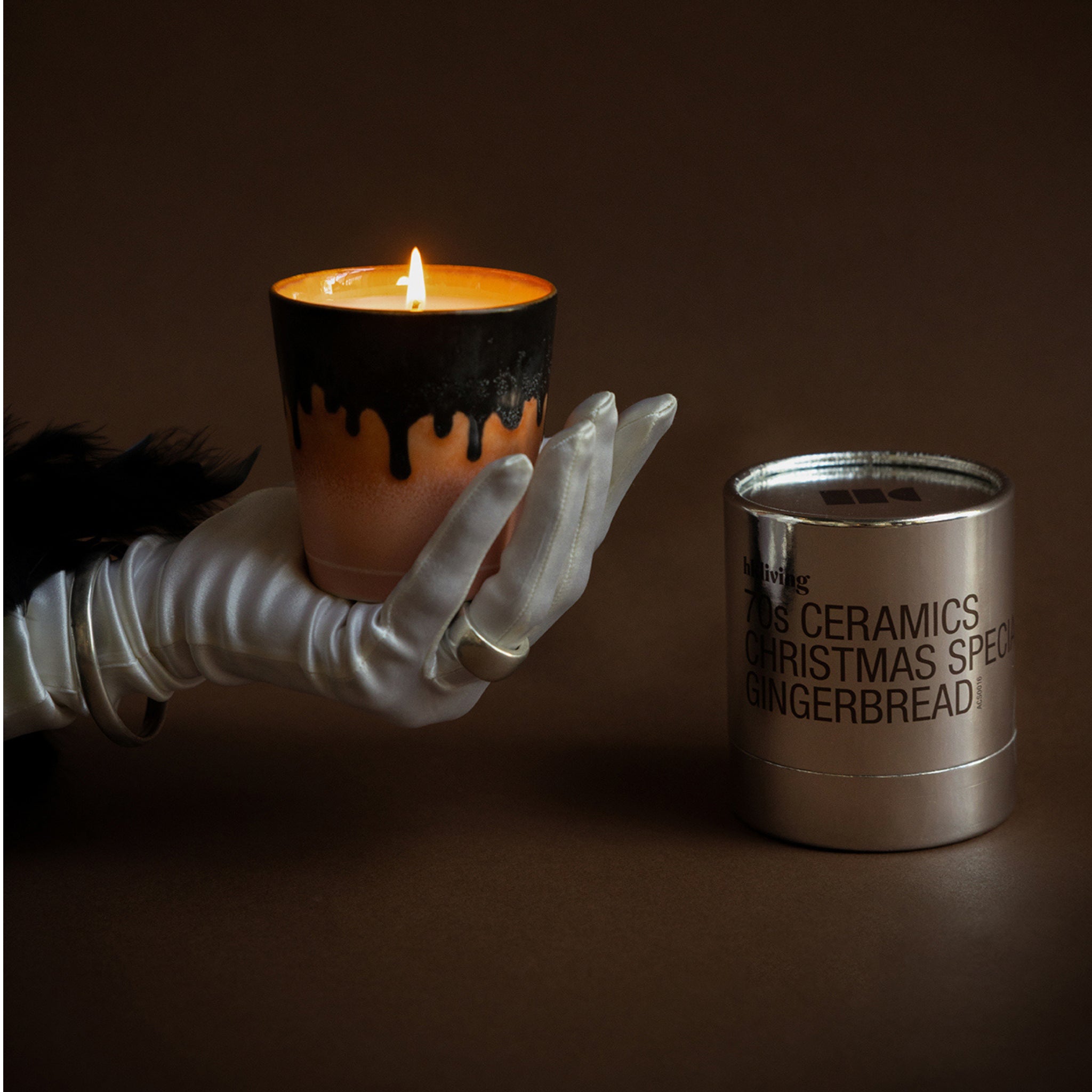 Een gehandschoende hand houdt een brandende kaars vast in een bruine keramieken houder met zwarte druppels. Ernaast staat een HK Living Christmas Special Geurkaars Gingerbread kaars in een metalen houder. De achtergrond is donkerbruin.