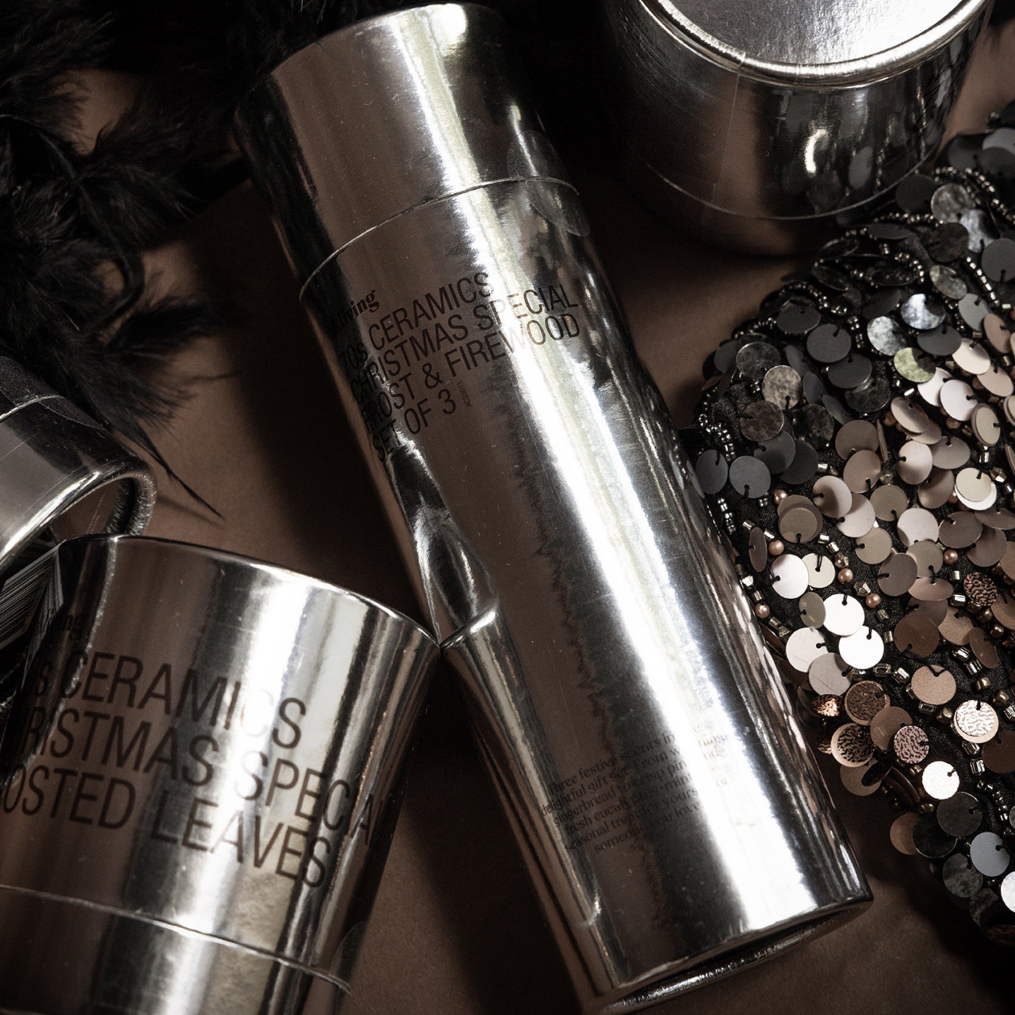 De HK Living Kerstspecial Geurkaarsen Frost & Firewood 3 st. in zilver metallic potjes met zwarte tekst worden getoond naast zwarte veren en een pailletten accessoire-een stijlvol kerstcadeau of om je favoriete geurkaarsen te presenteren.