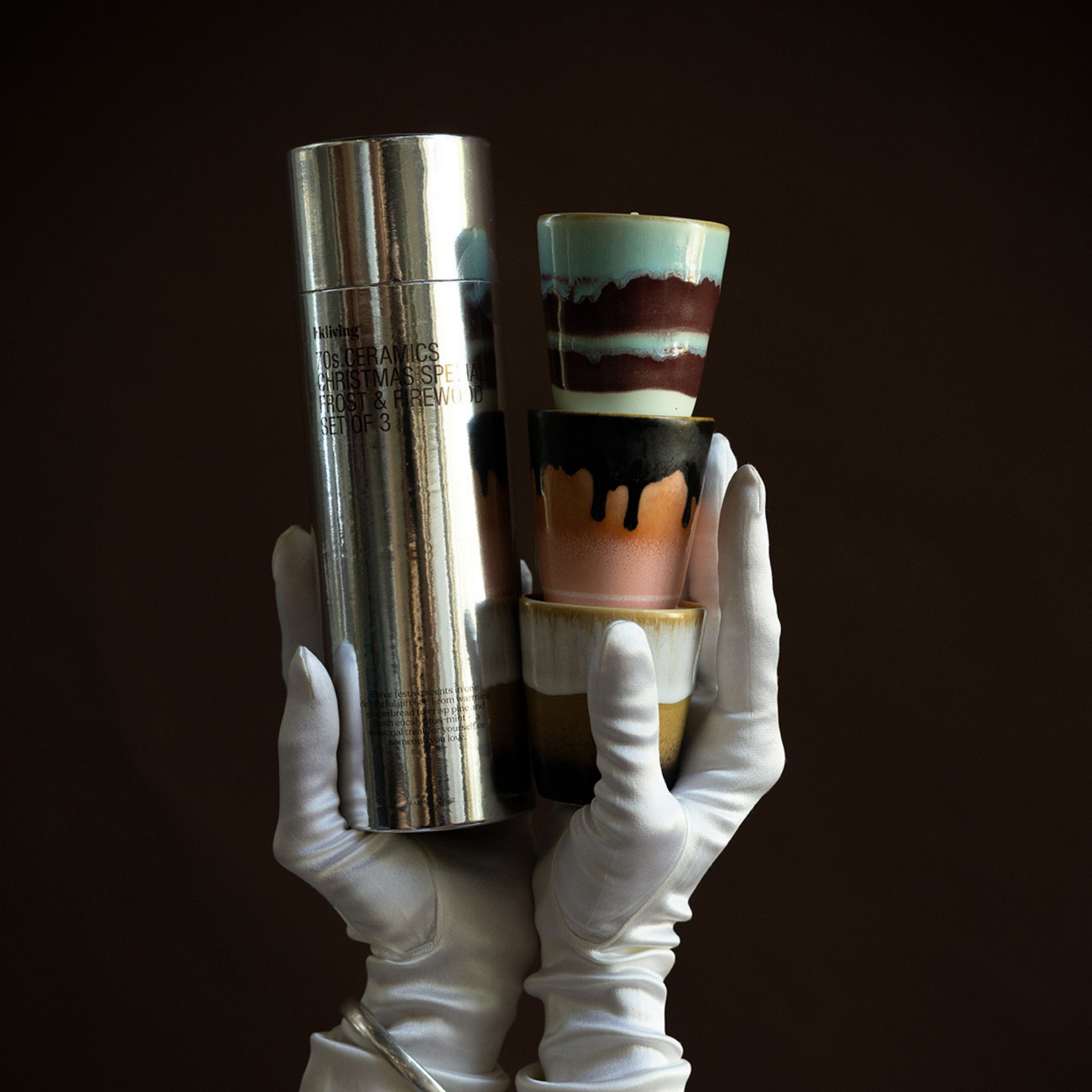 Een paar gehandschoende handen houden een glanzende zilveren bus vast naast een stapel HK Living Christmas Special Geurkaarsen Frost & Firewood 3 st. kopjes, met kleurrijke 70s Ceramics glazuurpatronen, tegen een donkere achtergrond.