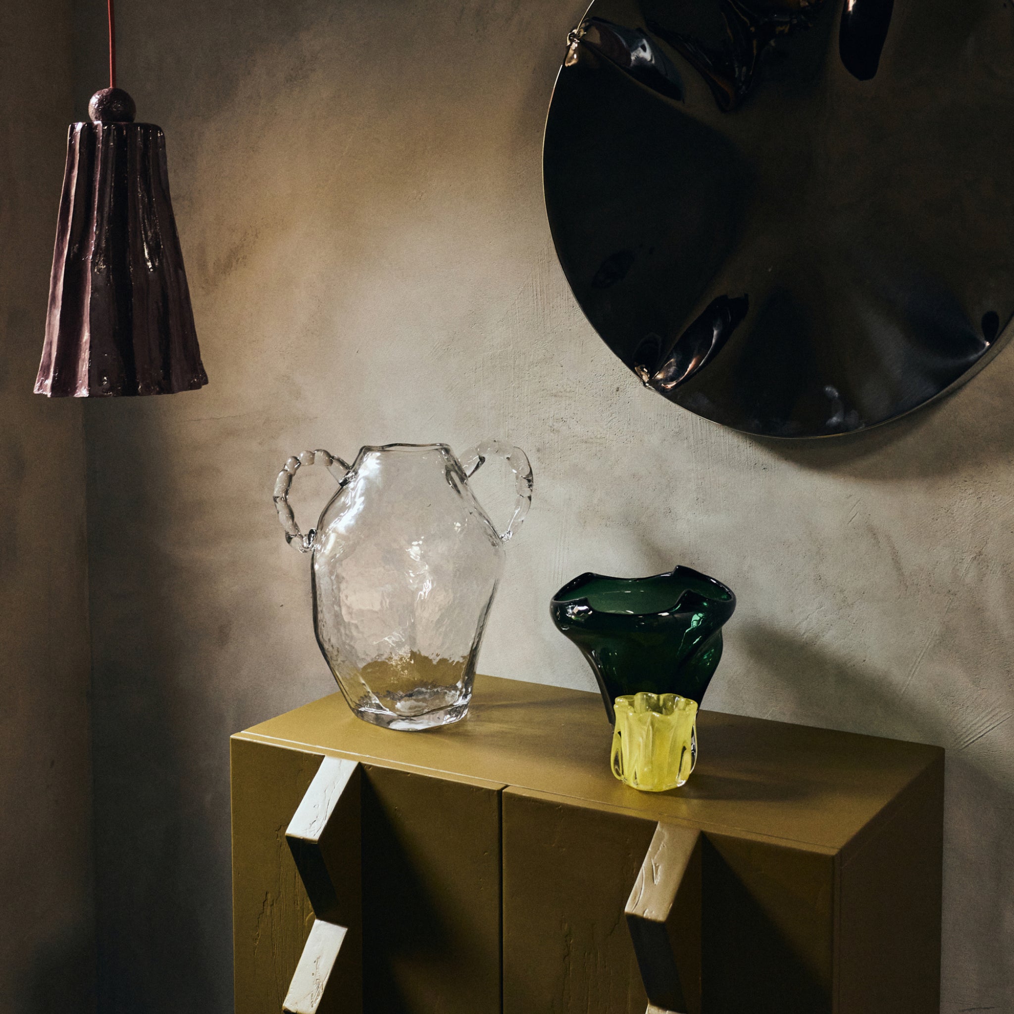 Een stilleven met een gestructureerde vaas van helder glas, een HK Living Coralla Theelichthouder Lime kom op een mosterdkleurige kast, een kastanjebruine hanglamp en een grote zwarte wandsculptuur tegen een grijze muur.