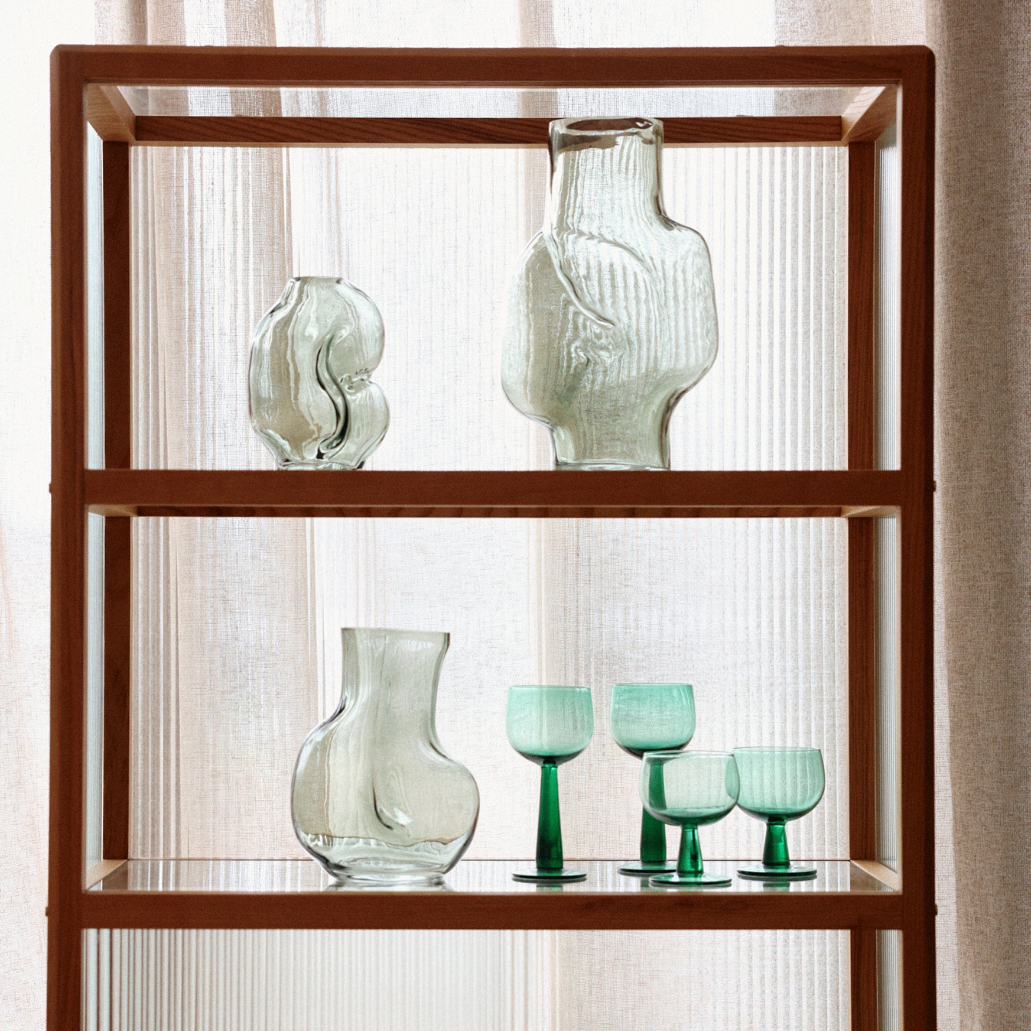 Een houten plank toont een HK Living Gleam Vaas Olijfgroen H22 op de bovenste en middelste etage, en vier glazen bekers met groene steel op de onderste plank, met gordijnen op de achtergrond.