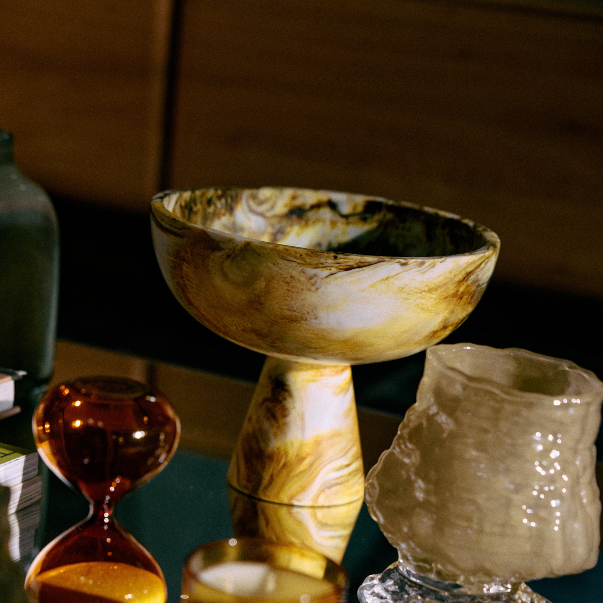 De HK Living gemarmerde Kom op Voet Oker D23 staat op een glazen tafel met een bruine zandloper, glazen vaas met structuur en gele kaarsenhouder - een ideaal decoratief object van HK Living voor elke stijlvolle ruimte.