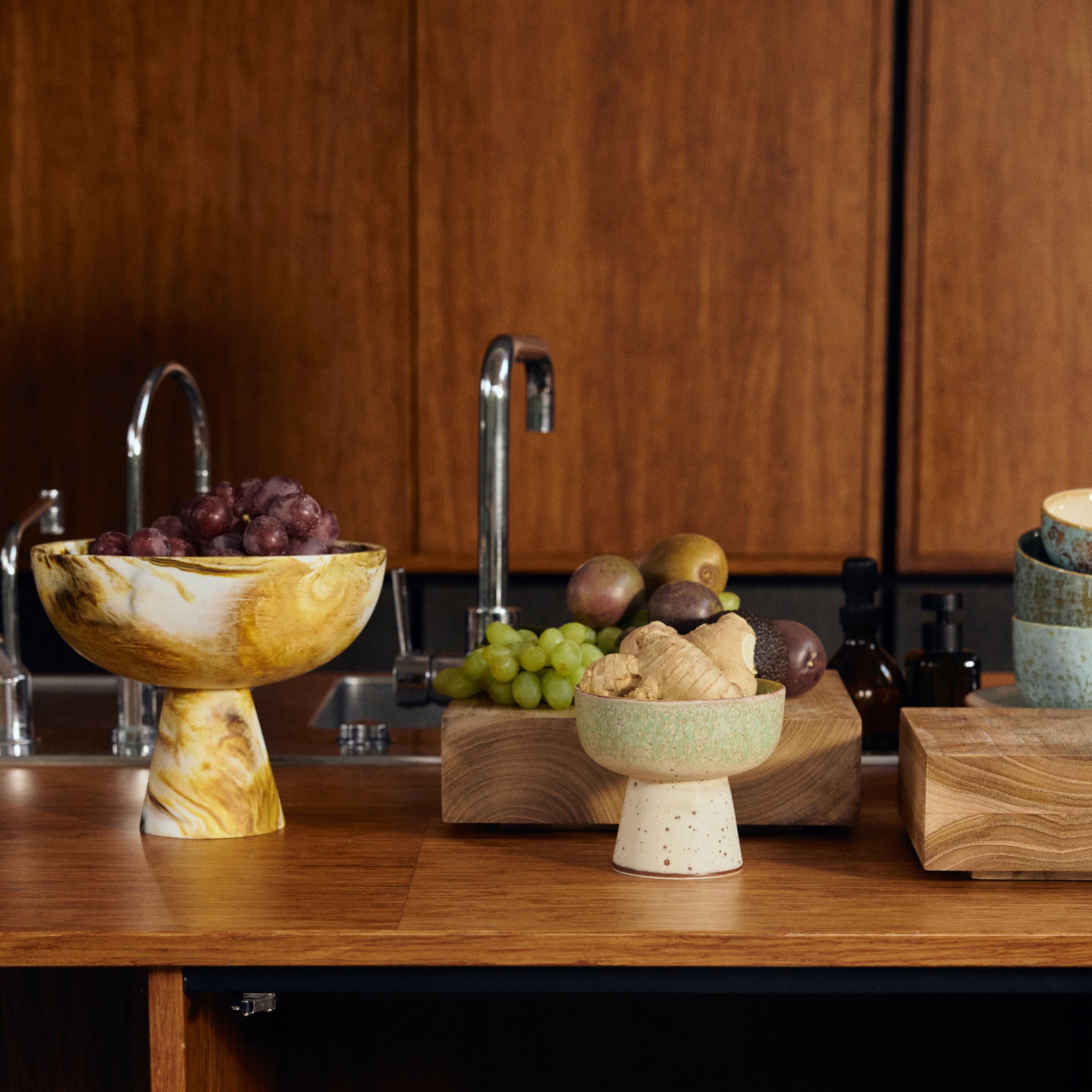 Op het aanrecht staat een HK Living gemarmerde Kom op Voet Oker D23 gevuld met rode druiven, naast gember, groene druiven, kiwi en bruin fruit op houten planken. Een gootsteen en bruine kastjes zijn zichtbaar op de achtergrond.
