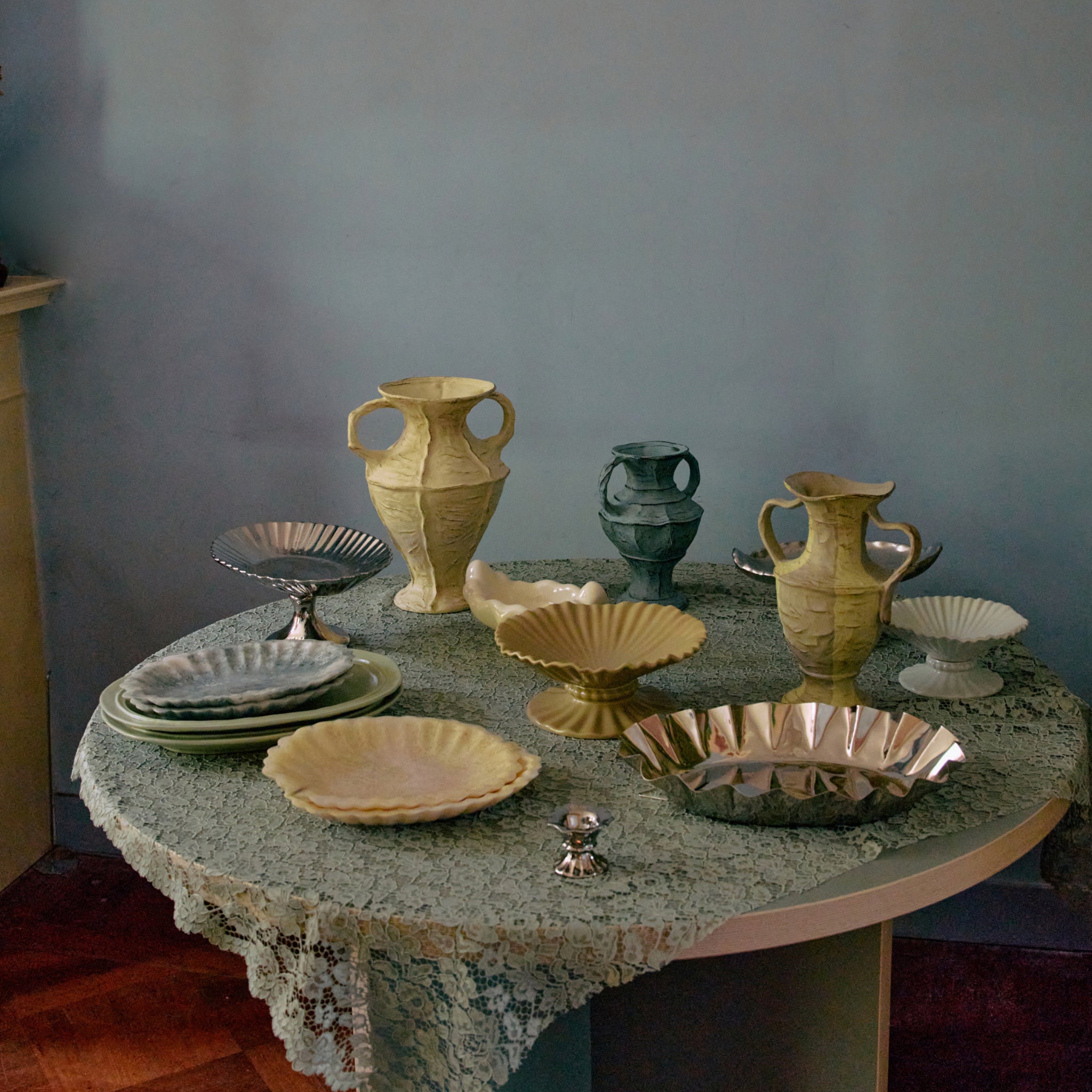 Op een ronde tafel met een kanten kleed staan keramische en metalen vazen, borden, schalen en de HK Living New Classics Resin Schaal Lichtblauw-allemaal netjes gerangschikt tegen een lichtblauwe muur.