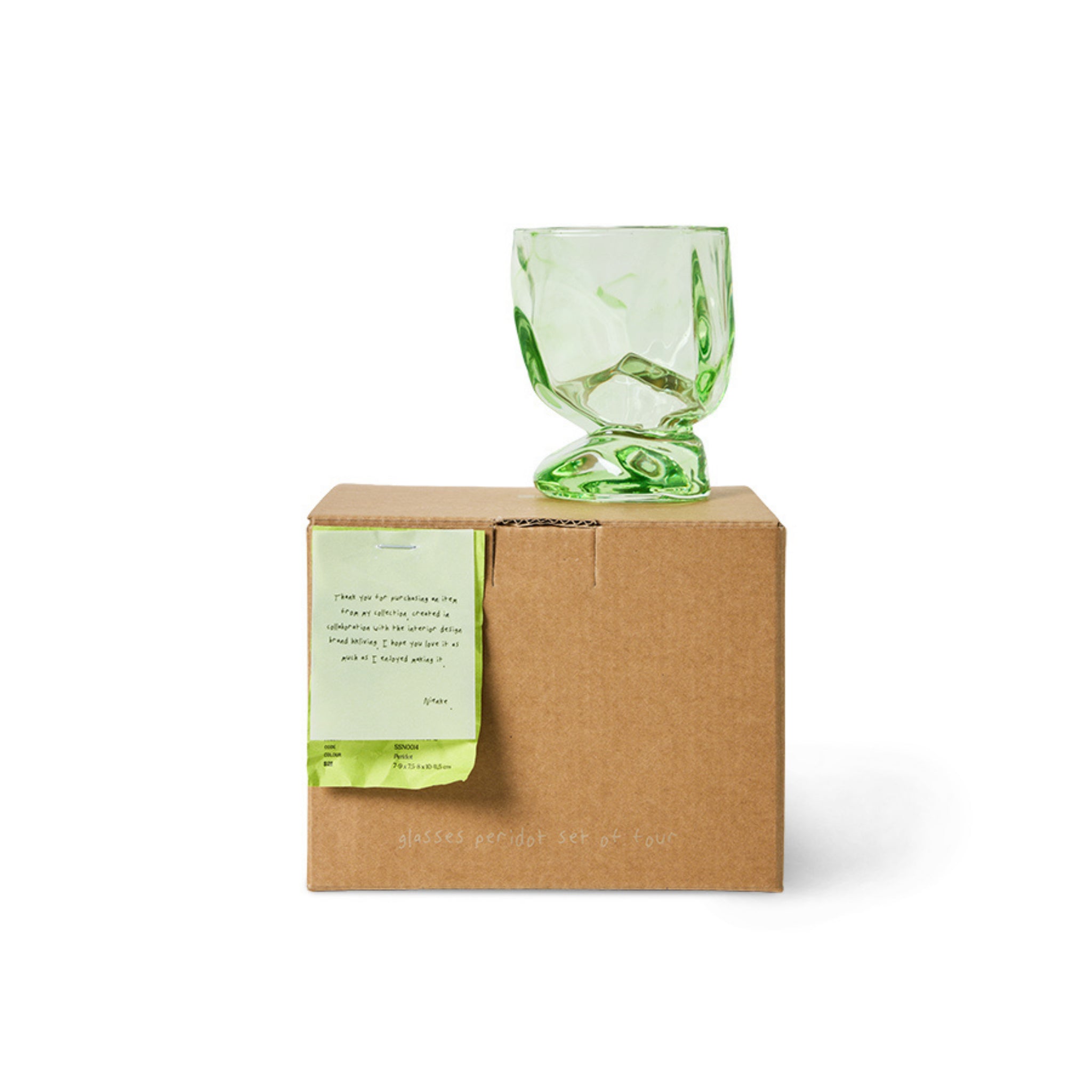 Een set van vier HK Living Nienke Sikkema Wijnglas Peridot Groen glazen zit bovenop een bruine kartonnen doos met een lichtgroen briefje aan de voorkant, alles tegen een witte achtergrond.