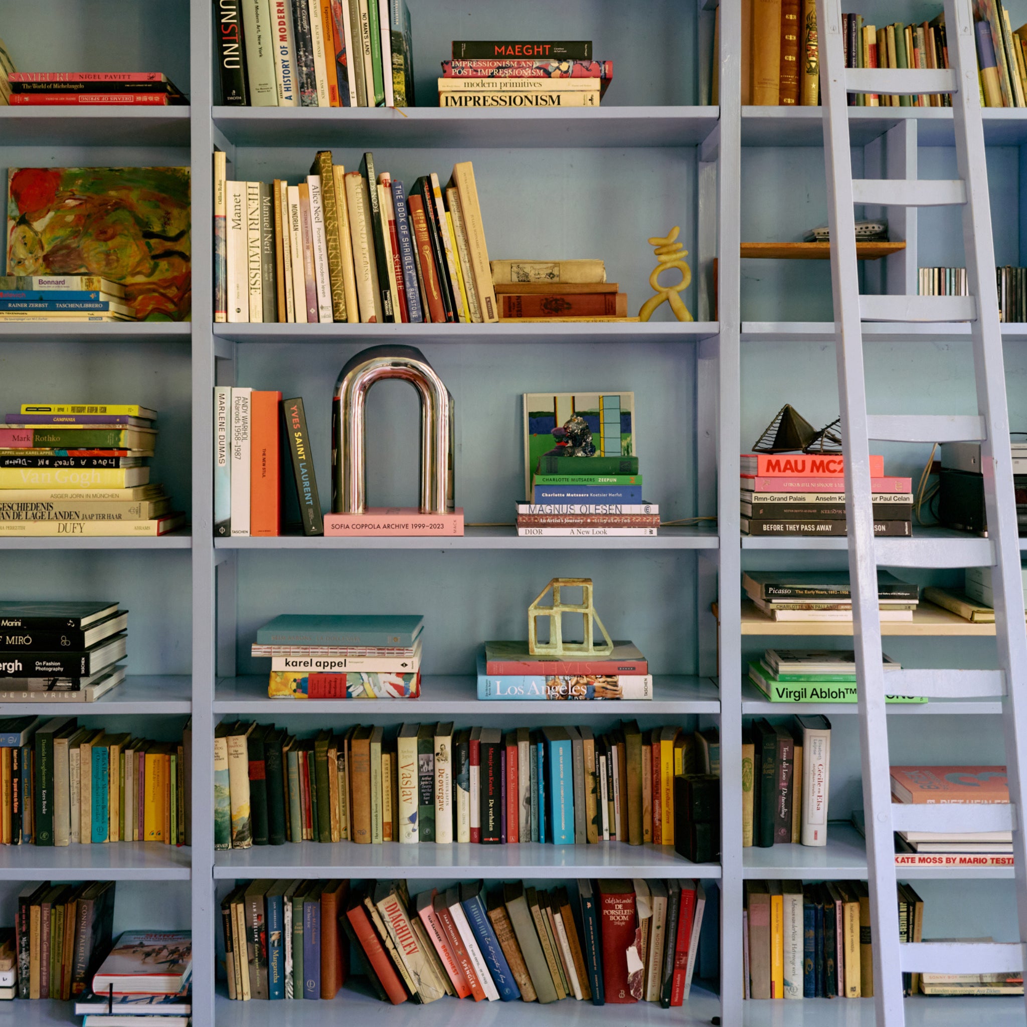 Een boekenplank met boeken in alle maten en kleuren, aangevuld met decoratief HK Living Paper Object: Naar Constructivisme en kunstwerken. Een rollende ladder leunt vlakbij en draagt bij aan de stijlvolle, eclectische sfeer van dit moderne interieur.