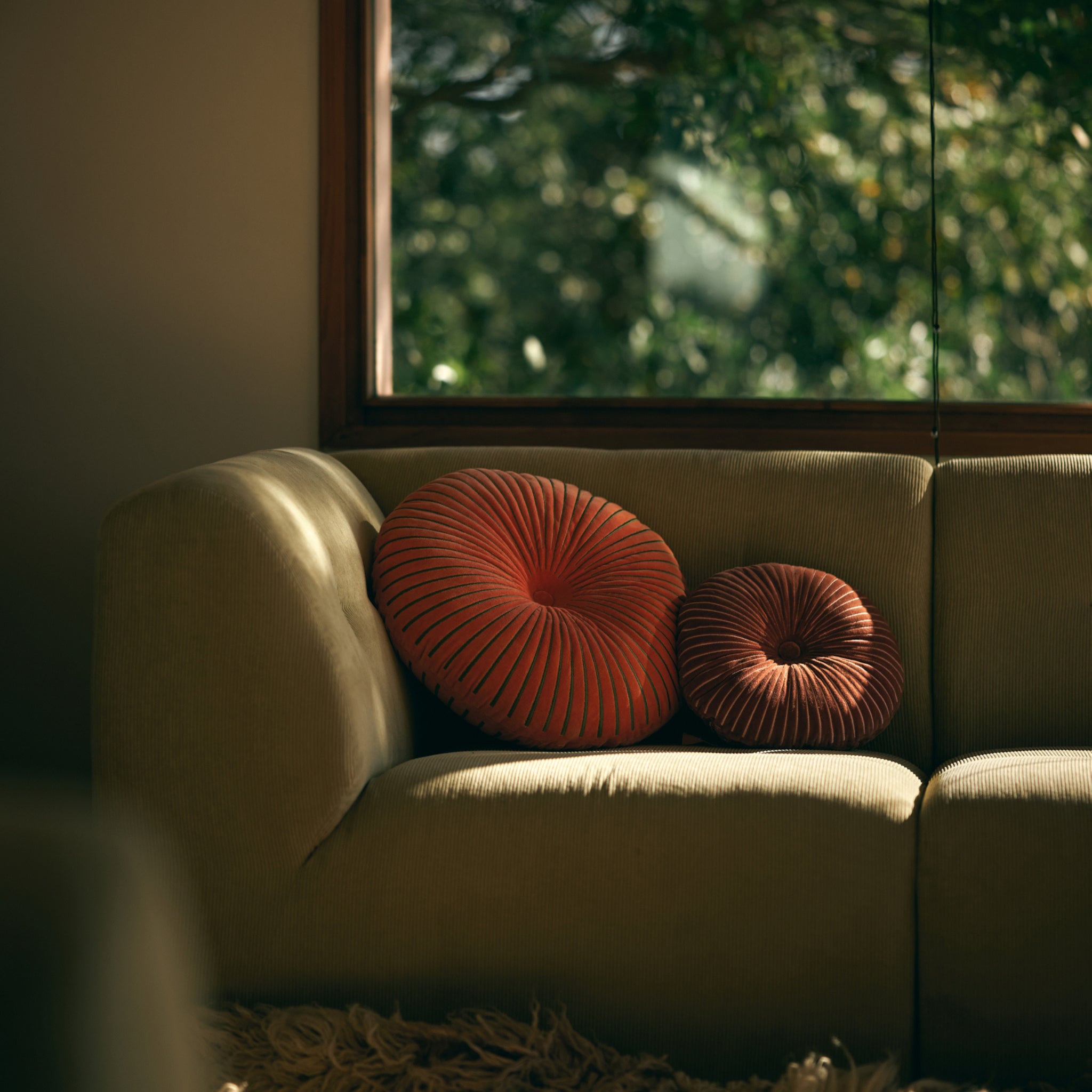 Twee HK Living Ripple Rond Kussen Sienna D50 kussens liggen op een beige bank bij een zonovergoten raam met uitzicht op weelderig groene bomen, waardoor de ruimte wordt gevuld met warm, natuurlijk licht.